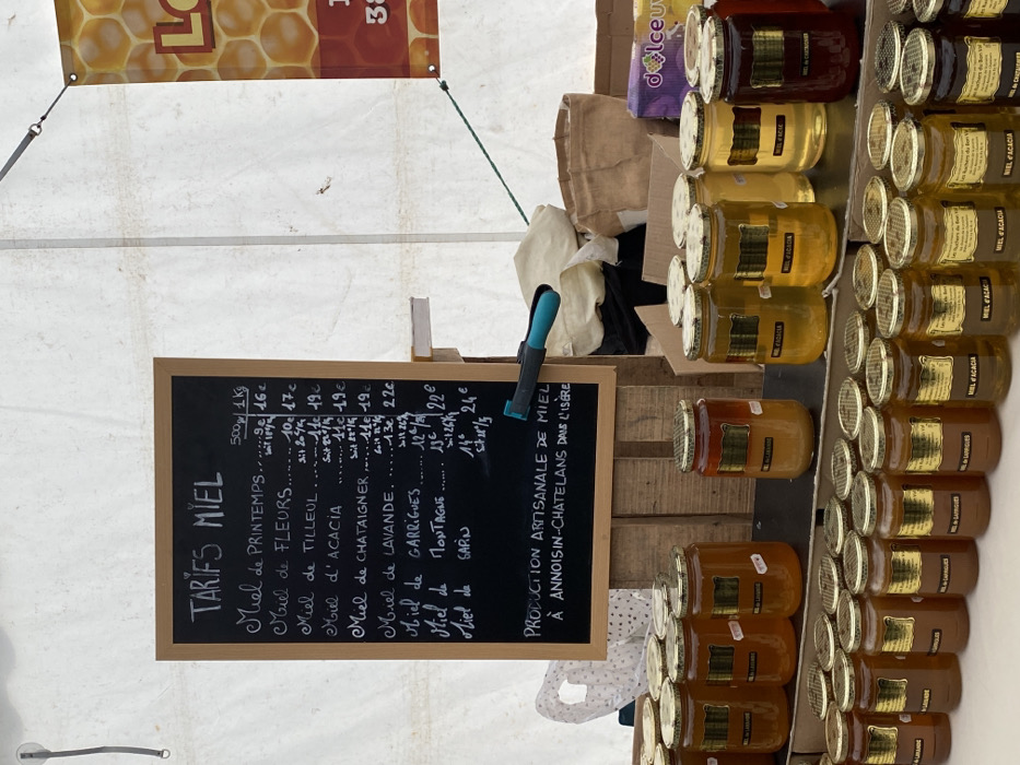 The image displays a honey market stall in Abondance, France, set up beneath a white canvas tent canopy. A prominent, vertically oriented blackboard sign, framed in wood, occupies the upper left-center, titled "TARIFS MIEL" (Honey Prices). The sign lists various honey types in white chalk, including "Miel de Printemps," "Miel de Fleurs," "Miel de Tilleul," "Miel d'Acacia," "Miel de Châtaigner," "Miel de Lavande," "Miel de Garrigues," "Miel de Montagne," and "Miel de Sapin," with prices provided for both 500g and 1kg quantities. Below the price list, the sign states "PRODUCTION ARTISANALE DE MIEL À ANNOISIN-CHATELANS DANS L'ISÈRE." A blue-handled clamp is attached to the right edge of the blackboard's support structure.
Below the sign, a tiered display table holds numerous uniform glass jars filled with honey. Each jar features a metallic golden screw-on lid and a rectangular golden label. Honey colors within the jars vary from light golden-yellow to rich amber and dark reddish-brown, reflecting different varieties such as "Miel de Lavande," "Miel d'Acacia," "Miel de Garrigues," and "Miel de Chataigner," as indicated on some labels. The display structure appears to be constructed from wooden crates or planks.
The background consists of the white canvas tent material, showing tension ropes and grommets. To the upper right, a portion of a banner with a honeycomb pattern and partially visible text, including "LO" and "38," is affixed to the tent wall. Behind the honey display, miscellaneous items are visible, including a crumpled light-colored cloth, a brown burlap sack, a cardboard box, and a purple package labeled "dolceuv." No individuals are present within the frame.