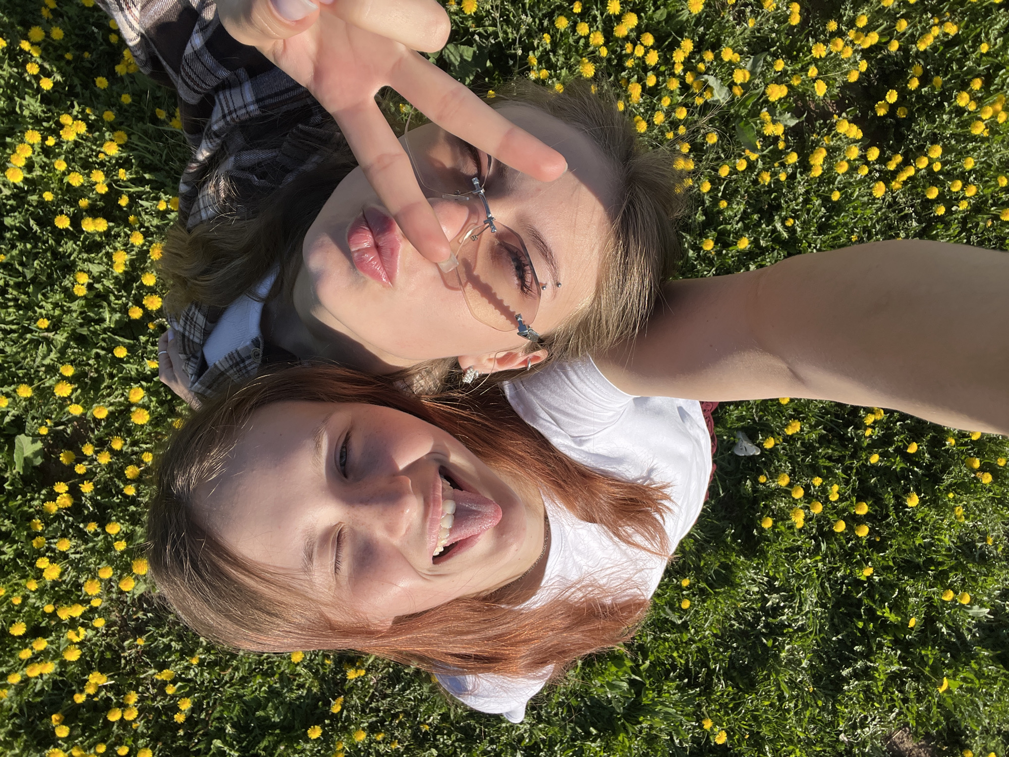 Two young women are lying on a grassy field covered in yellow dandelions. One woman is wearing glasses and making a peace sign. The other woman has her tongue sticking out. They appear to be taking a selfie. The location is not identifiable from the image.