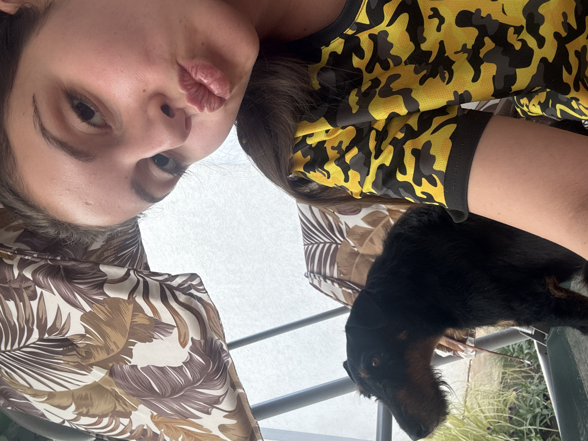 A young woman with dark hair is taking a selfie outdoors, her face close to the camera, puckering her lips. She is wearing a bright yellow shirt with a black camouflage pattern. To her left and slightly behind her, a black dog with brown markings is visible, looking to the left. They are sitting on a swing set with patterned cushions. The background includes foliage and a white wall or sky. The location is Bulgarovo, Bulgaria.