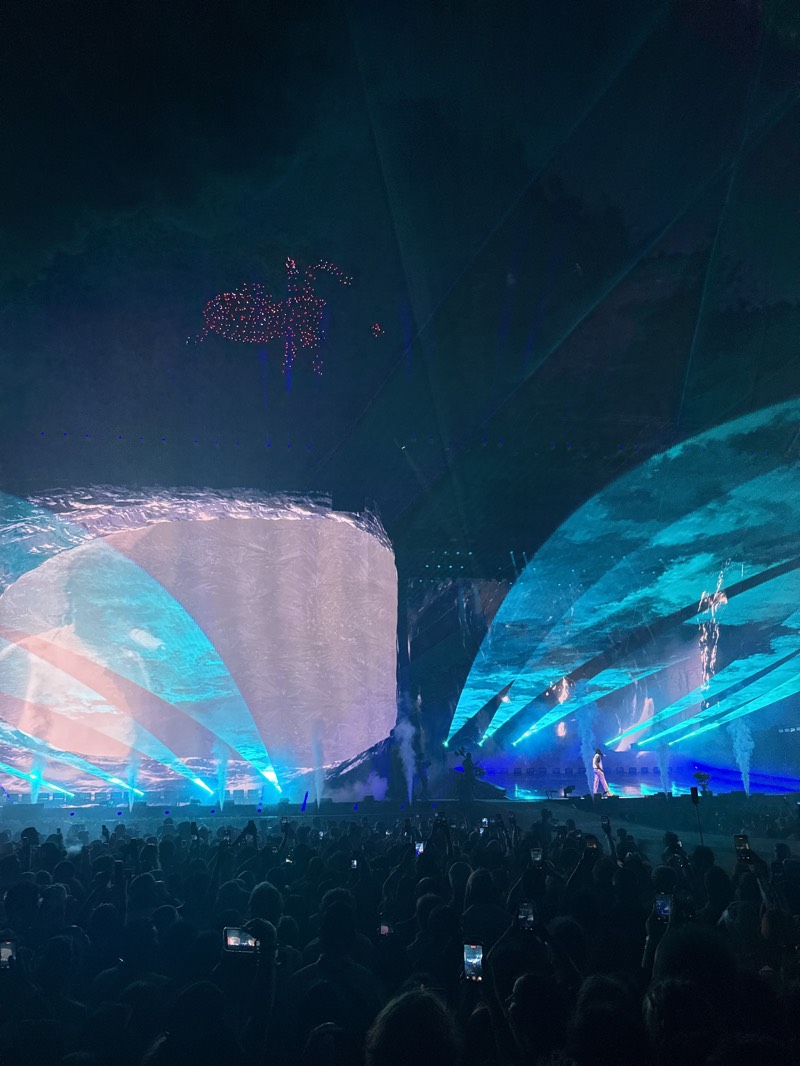 The image depicts a large crowd attending a concert in Indio, United States. A stage is set up with a large screen displaying abstract imagery in shades of blue and white. Laser beams of blue light emanate from the stage, illuminating the performer in the center. The audience is densely packed, with many individuals holding up phones to capture the performance. Above the stage, a formation of lights, possibly drones, is arranged in the shape of a figure, resembling a horse rider. The atmosphere is dark, with the stage lights and drone formation providing the primary illumination.