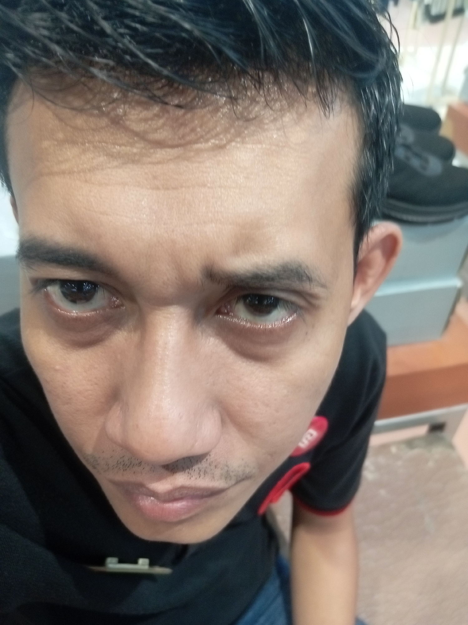 A close-up, slightly angled, selfie-style photograph shows the upper body and face of a man with dark hair and fair skin. The man is wearing a black t-shirt with red trim and a logo on the sleeve. His expression is serious, with furrowed brows and his eyes looking directly at the camera. The background is out of focus, but appears to be an indoor setting with shelves and what look like shoes on a shelf. The lighting is bright and direct, creating some glare on the man's forehead and nose. There is no visible evidence to indicate the specific location within Batam, Indonesia.