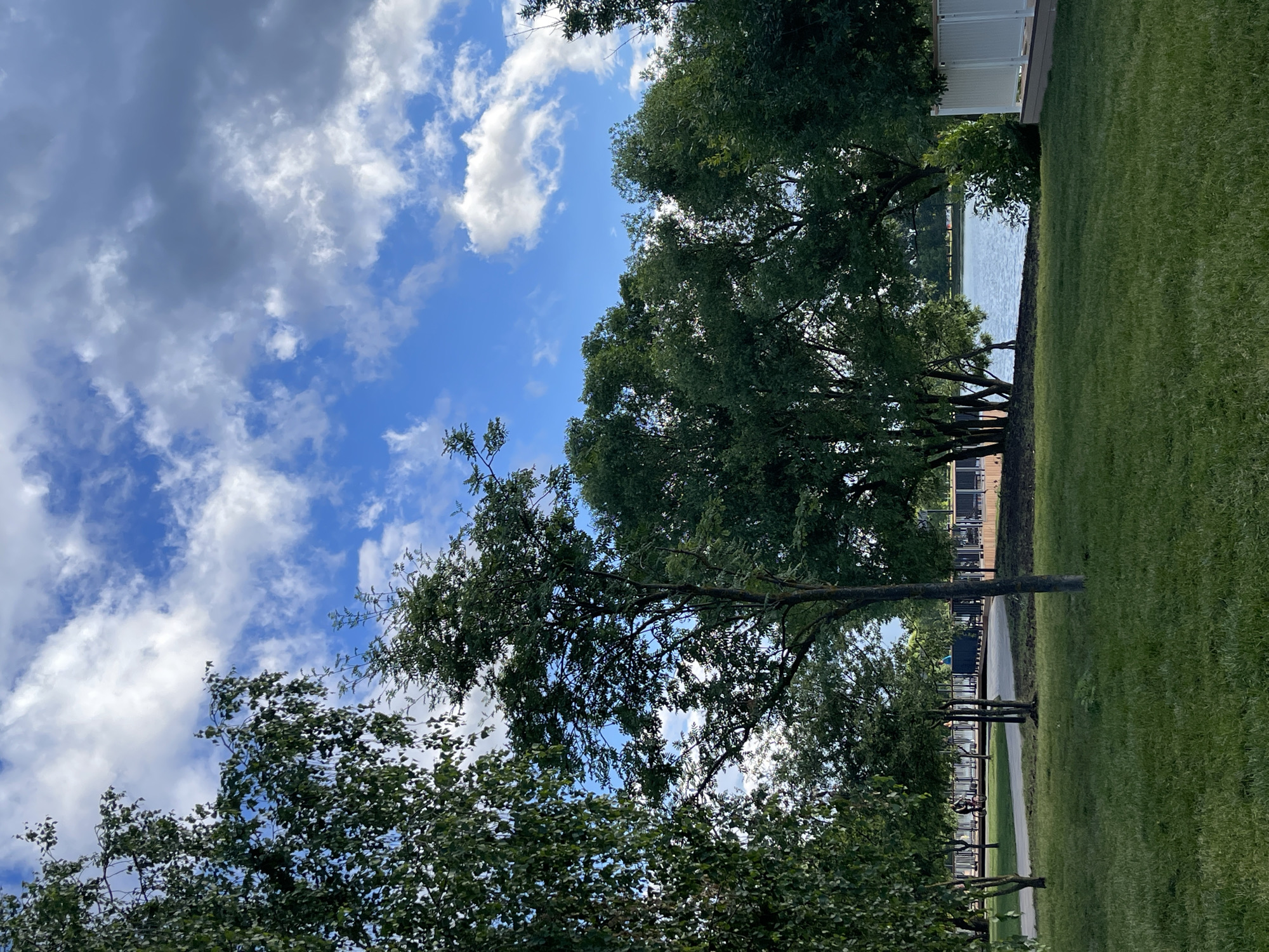 The image depicts a park area with a body of water in the background. Lush green grass covers the foreground, with several trees of varying sizes scattered throughout. In the middle ground, a paved path winds through the park, with a few individuals visible. One person is riding a bicycle, while others are walking. Beyond the path, a low wooden fence or platform is visible along the water's edge. The body of water is calm, with ripples on its surface. In the distance, more trees and structures can be seen. The sky above is a mix of blue sky and white and grey clouds. The scene suggests a recreational area within a city, likely a park or waterfront promenade. The location is identified as Klyuch Monument Park in Moscow, Russia.
