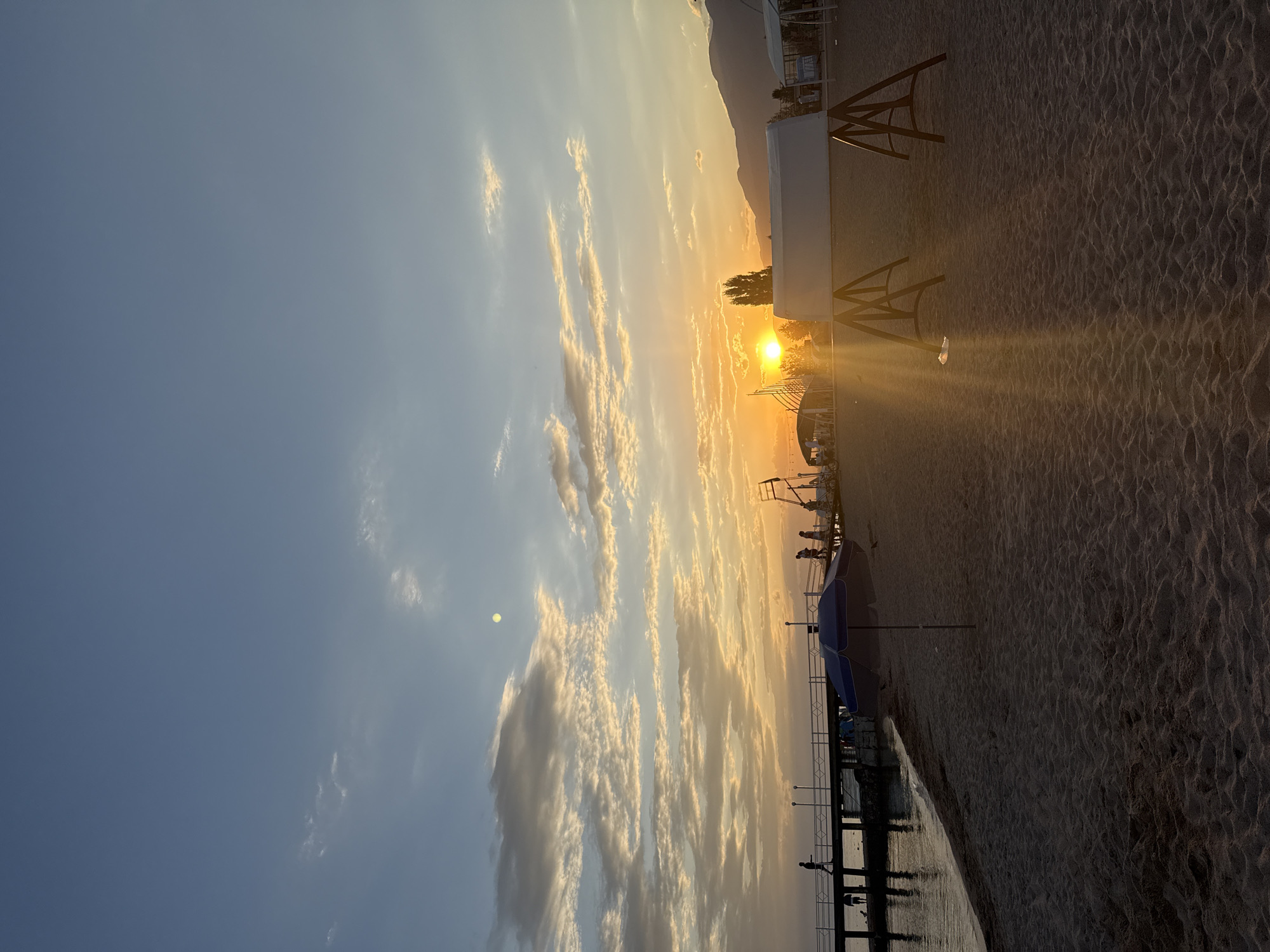 An outdoor, vertical, wide shot captures a sunset on a sandy beach in Sary-Oy, Kyrgyzstan. The sky is a gradient of blue and orange, with clouds scattered throughout. The sun, a bright orange orb, is partially obscured by clouds and low-lying trees. A wooden pier juts out into the water on the left side of the frame, with several silhouetted figures walking along it. On the beach, a blue and white umbrella stands near the water's edge, and a white tent-like structure with wooden supports is in the foreground. Mountains are visible in the distance, silhouetted against the colorful sky. The sand is dark and appears to be wet near the water. The overall mood is serene and peaceful, with a warm glow from the setting sun.