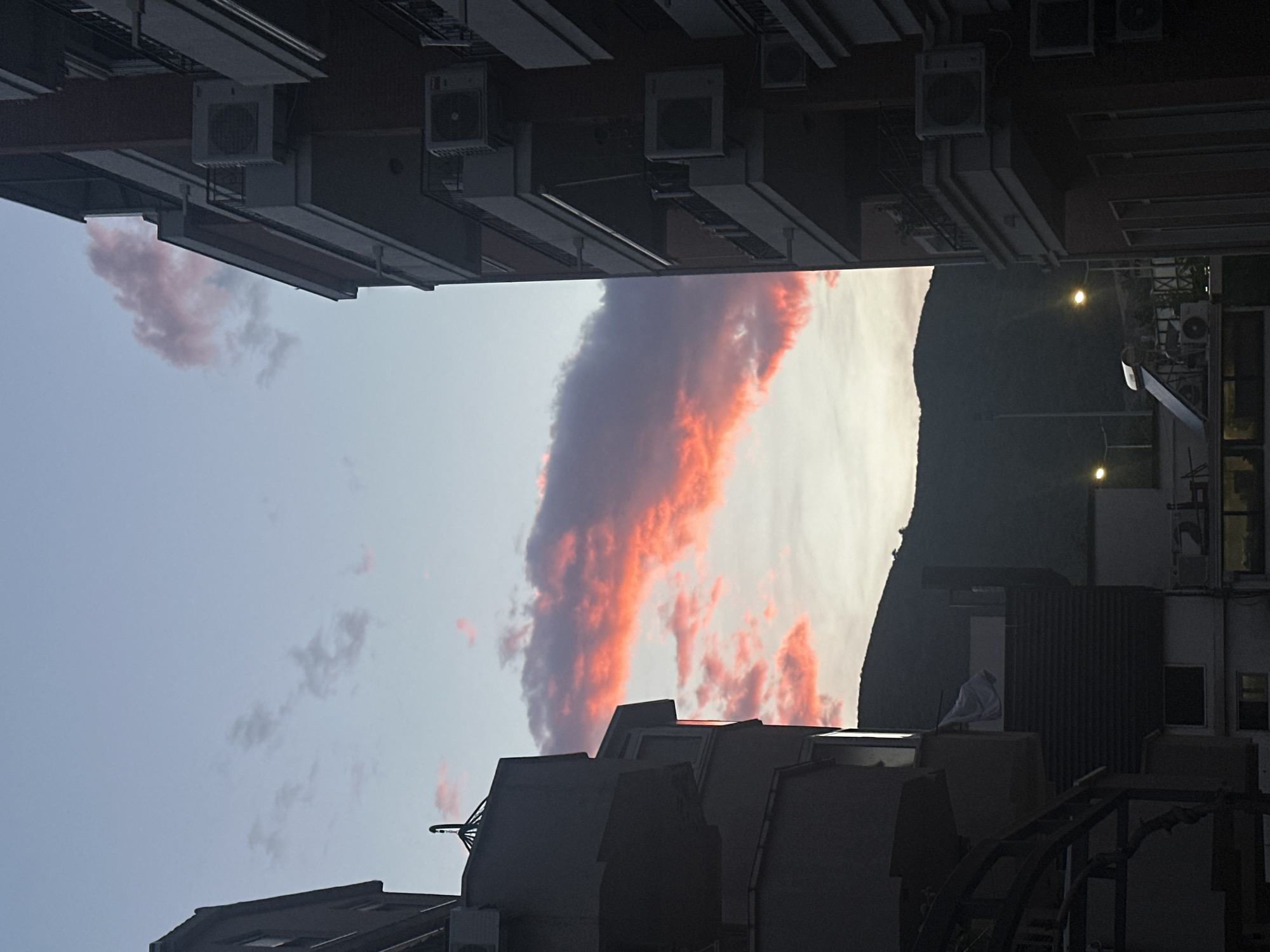 The image, taken in a likely urban environment, captures a view upwards between buildings towards a sky with scattered clouds. The buildings on the left and bottom are silhouetted against the sky, displaying architectural details such as air conditioning units attached to the building exteriors.
The sky features clouds, some of which are illuminated in hues of pink and orange, suggesting either sunrise or sunset. A dark landform is visible on the right side, possibly a hill or mountain. Streetlights are visible on the right side of the image.
The overall impression is one of a densely built urban area with a view to the sky at dawn or dusk. Location cannot be ascertained.