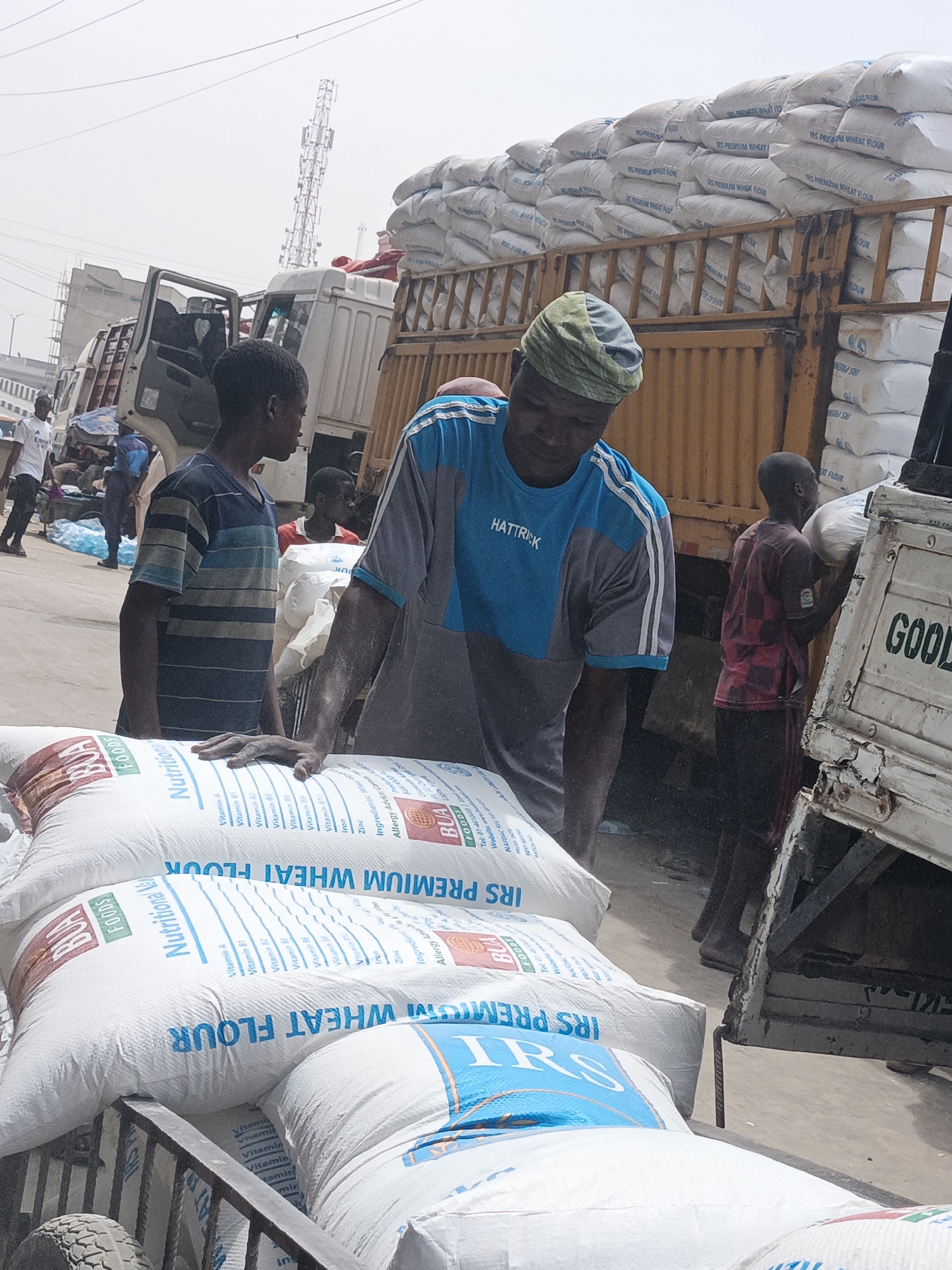 In Fagge C, Nigeria, a bustling street scene unfolds with several people involved in the unloading of goods. At the forefront, a man wearing a gray and blue "HATTRICK" shirt and a head covering rests his hand on stacks of "IRS PREMIUM WHEAT FLOUR" bags loaded onto a hand truck or cart. Adjacent to him, a boy in a striped shirt stands looking at the stacks of flour. A younger boy is actively offloading bags from the back of a white truck, labeled with "GOOD" in large letters. A larger, yellow-painted truck is visible behind them, piled high with more bags of "IRS PREMIUM WHEAT FLOUR". Other figures are present in the background, along with vehicles and indistinct structures suggesting a busy commercial environment. Overhead power lines and a cell phone tower are also visible.