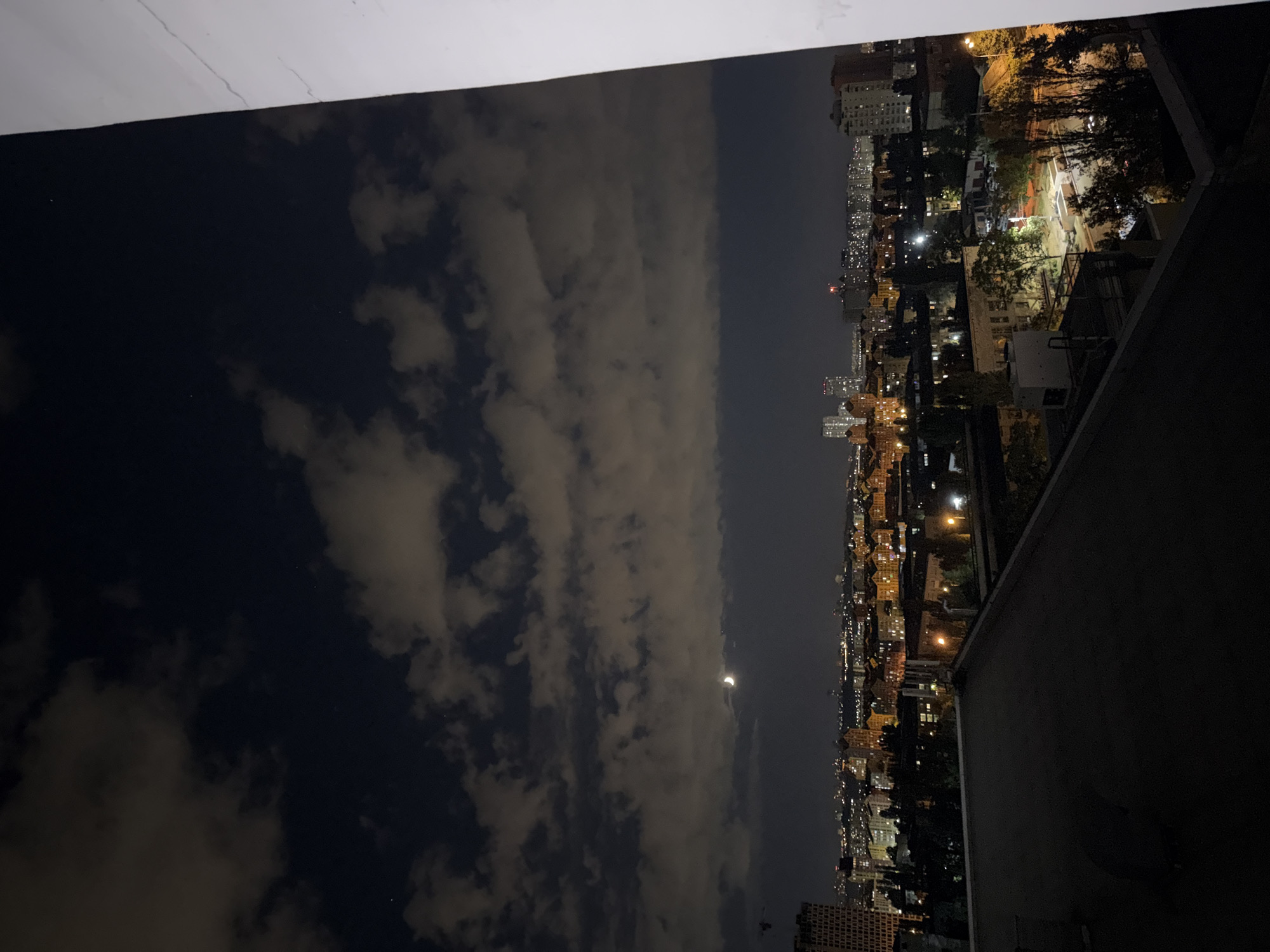 This is a nighttime photograph taken from a rooftop overlooking a densely populated urban area, possibly in Kyiv, Ukraine, given the architectural style of the buildings and the presence of a crescent moon. The foreground shows the dark surface of a rooftop, with a white wall visible on the right side. Beyond the rooftop, the cityscape unfolds with numerous buildings of varying heights, many illuminated by warm, artificial lights. Trees are scattered throughout the urban landscape, and streetlights cast a glow on the streets. In the sky, a crescent moon is visible amidst scattered clouds. The overall impression is one of a city at night, with the typical hum of urban life continuing under the celestial glow.