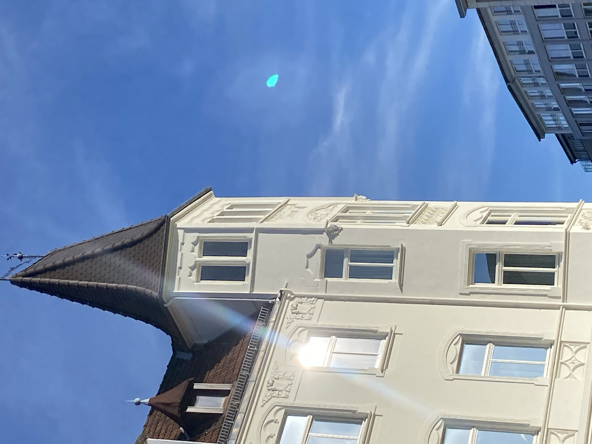 The image captures an architectural detail of a building in Bielefeld, Germany, under a clear blue sky with some wispy clouds. The main subject is the corner of a light-colored building with a distinctive conical dark-tiled roof topped by a spire. The building features several windows with white frames. Sunlight reflects brightly off some of the windows, creating a lens flare effect. To the right of this building, another modern building with a grey facade and numerous windows is visible, angled away from the viewer. In the sky, a small, bright blue oval-shaped object is present. The scene appears to be a daytime shot, likely in an urban setting, focusing on the architecture and the sky. There are no people visible in the image.
