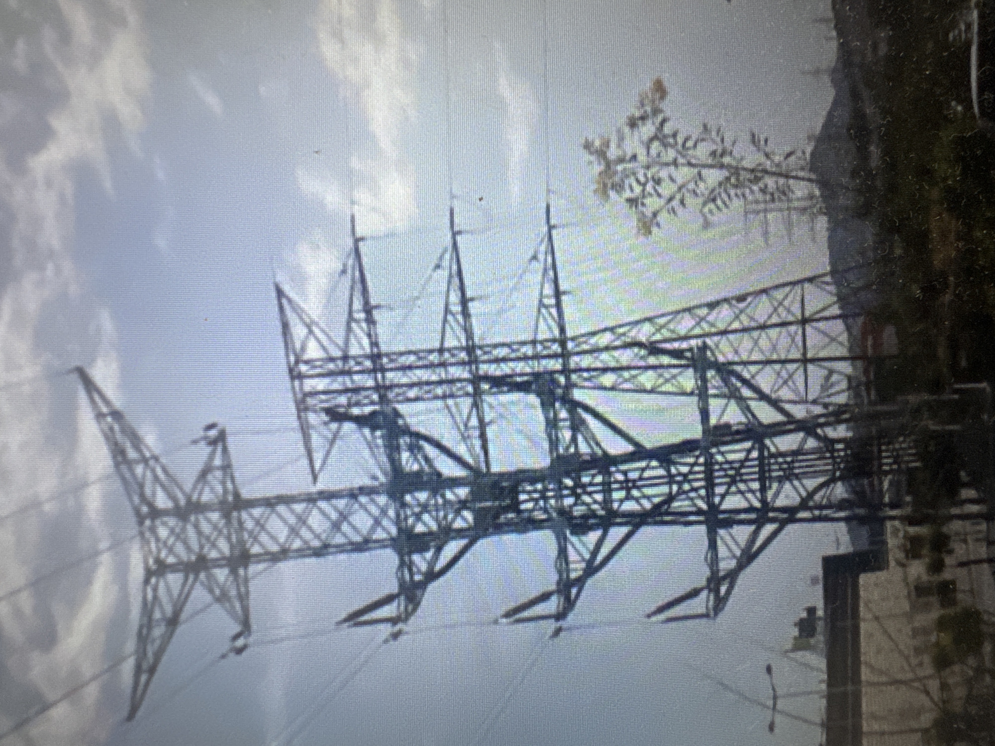 The image shows two large electricity pylons against a cloudy sky. The pylon on the left is closer to the viewer and appears to be taller. The pylon on the right is slightly behind and to the right of the first. Several electrical wires are strung between the pylons. In the foreground, there are some buildings and vegetation. The overall scene suggests an industrial or semi-urban environment. The presence of electrical pylons is a common feature in many locations, and without additional context or specific visual cues, it is not possible to definitively determine the location as the city of Murcia, Spain.