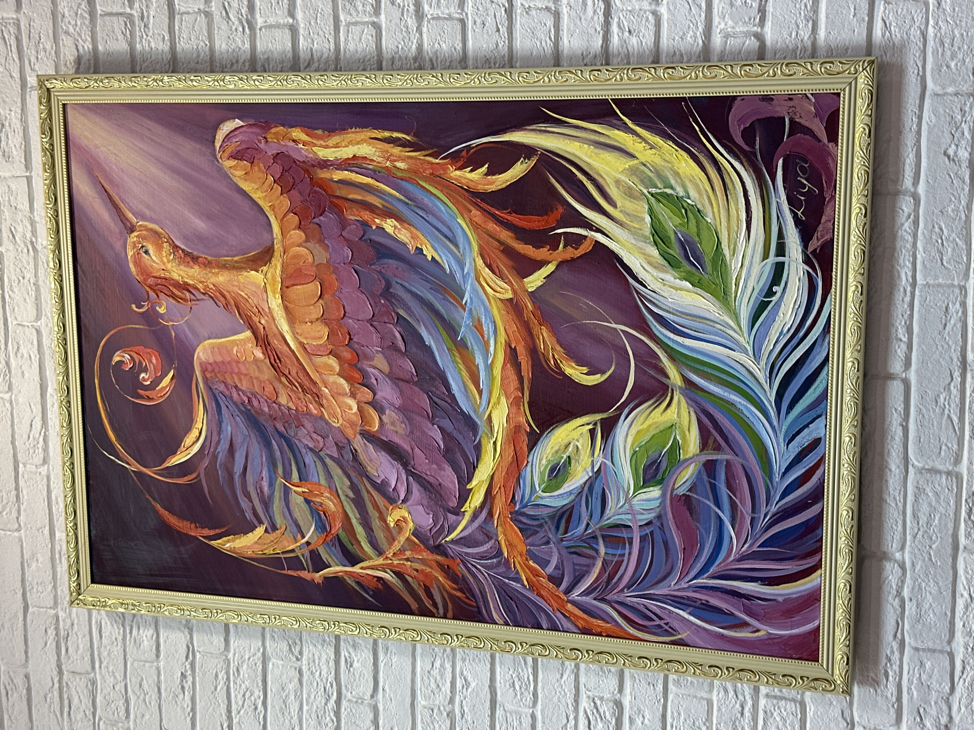 A framed painting hangs on a white brick wall. The painting depicts a phoenix-like creature with colorful feathers, primarily in shades of orange, yellow, purple, blue, and green. The background is a dark purple, with lighter shades creating a sense of light and shadow. The frame is ornate and gold-colored. There is a signature on the upper right side of the painting. The location is Unknown, Unknown.