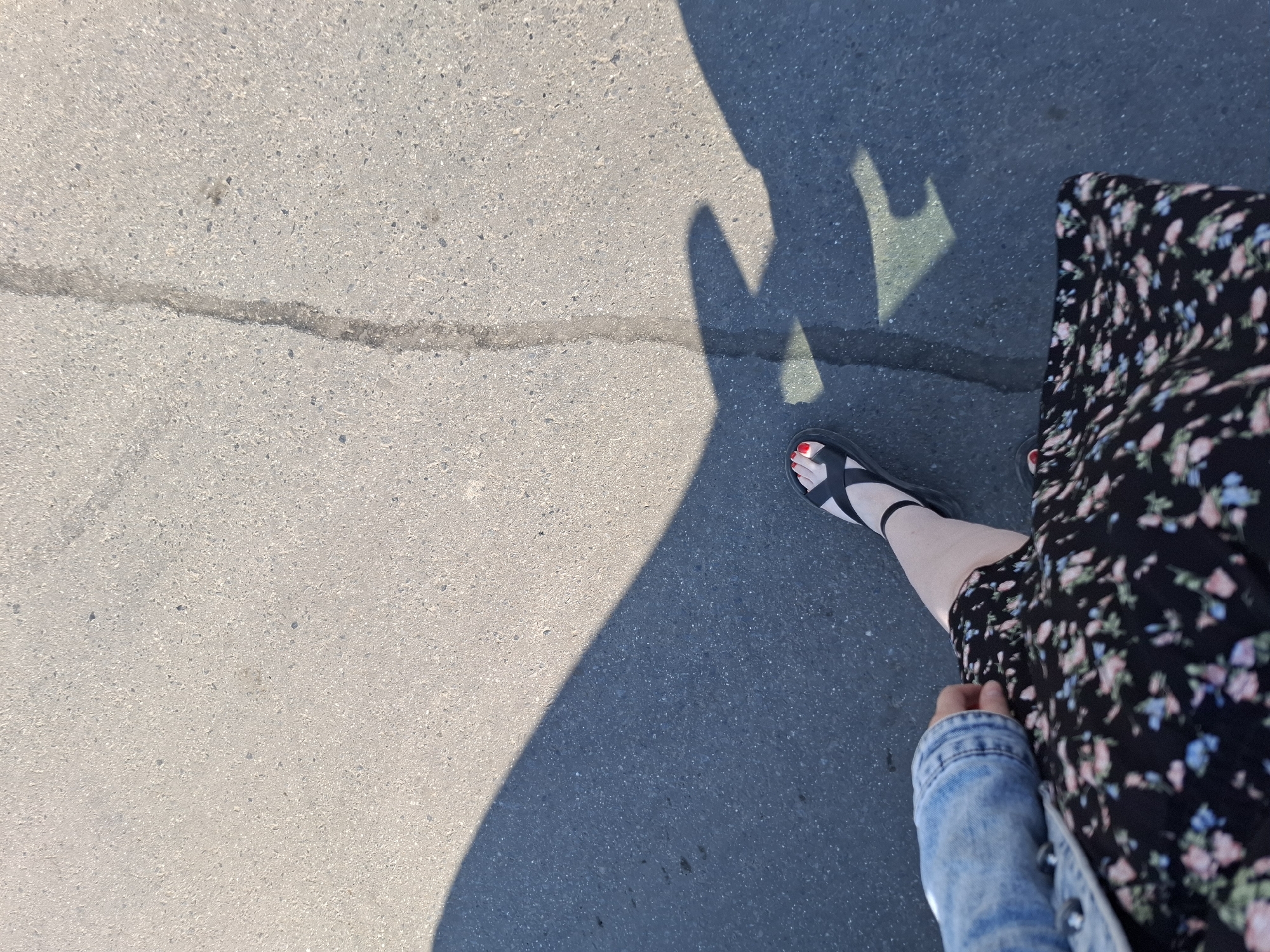A top-down perspective shows a person standing on a concrete surface. The person is wearing black sandals with red nail polish visible on their toes, a black dress with a floral pattern, and a blue denim jacket. The person's shadow is cast onto the concrete surface, appearing elongated and distorted due to the angle of the light source. The concrete surface has a visible crack running across it. The location appears to be a public space, possibly a sidewalk or a paved area, somewhere in Izhevsk, Russia.