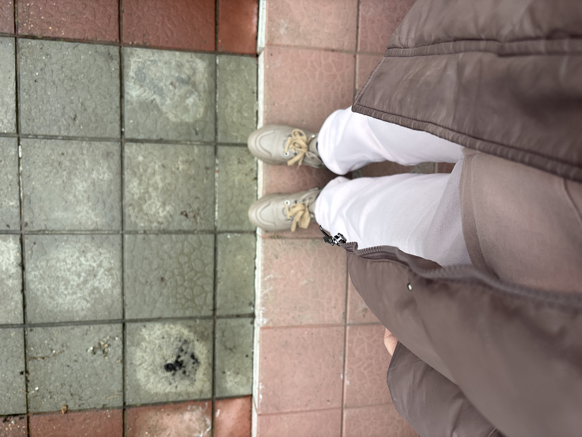 The image, taken from an overhead perspective, shows a person standing on a tiled surface. The tiles are a mix of grey and reddish-pink. The person is wearing white pants and light-colored sneakers with tan laces. A brown puffer jacket or vest is partially visible, obscuring part of the view. The location is identified as Shul'gino, Russia. The image mainly captures the person's footwear and the ground, with minimal surrounding context.