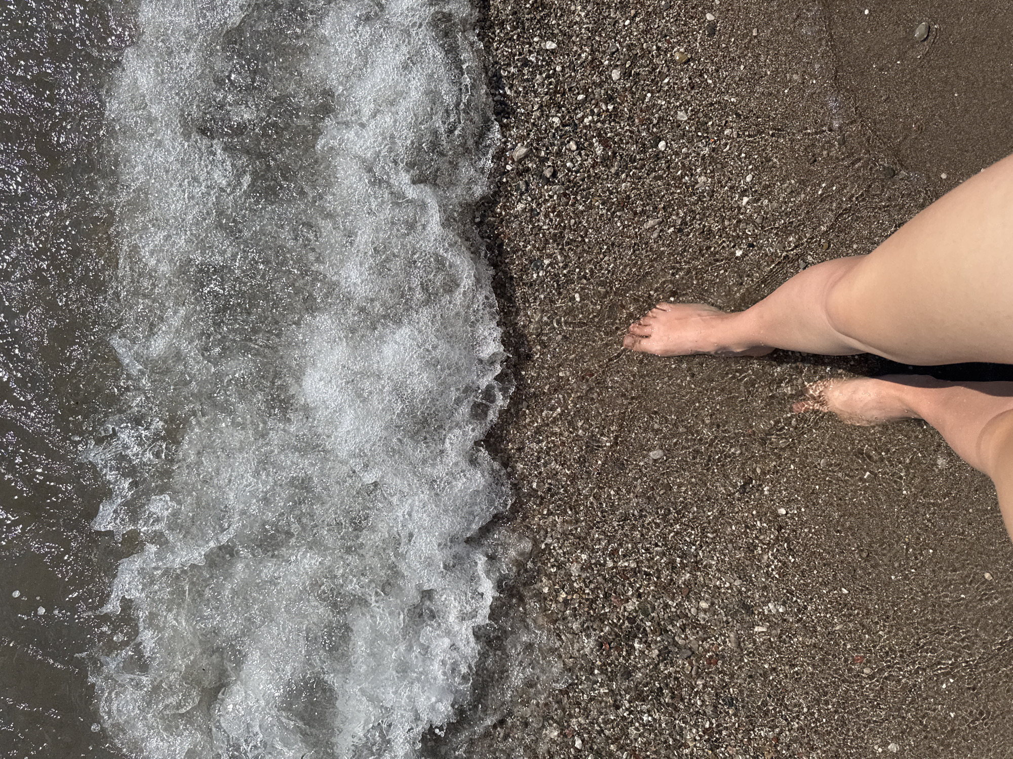 This image shows a top-down view of a person's bare feet standing at the edge of the sea on a pebbly beach. The water is clear and appears to be a shallow wave. The beach is covered in small, dark-colored pebbles. The scene is consistent with a location along the coast of Šušanj, Montenegro, a popular seaside resort town. The lighting suggests it is daytime, likely a sunny day.