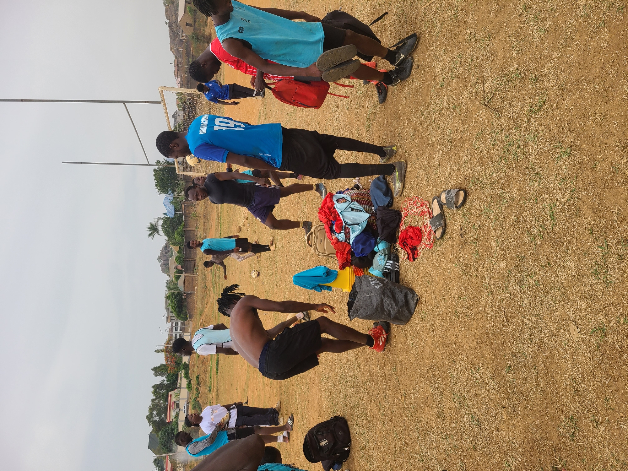 The image depicts a group of young men engaging in an apparent soccer practice in Barikin Jirga Summa, Nigeria. The setting is an open field covered mostly with dry grass and patches of bare earth, suggesting a semi-arid climate. In the background, there are low-rise buildings and trees, characteristic of a residential area in the town. The weather appears to be overcast.
Several men are seen stretching or preparing for a game. A soccer goal post is visible. Clothes and belongings, including bags and shoes, are scattered on the ground. The men are wearing a mix of sports attire, with some shirtless or wearing athletic jerseys. One person is identifiable by the number '19' on their blue shirt. Their actions indicate physical activity and preparation for a soccer game.