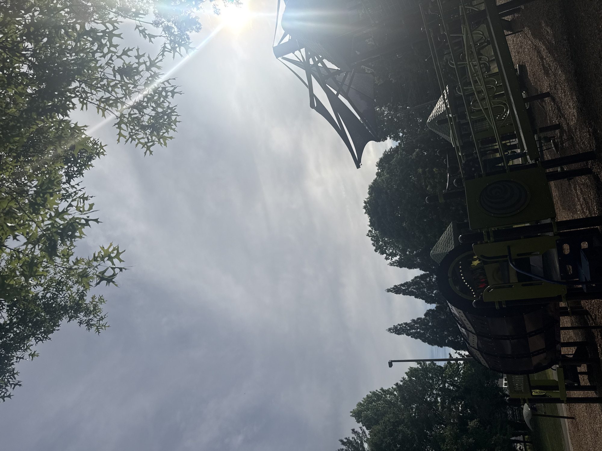 The image captures a scene at a playground in Mountain Park, Georgia, USA. The focus is on playground equipment, including a tall climbing structure with a dark-colored fabric shade and a cylindrical structure with windows. Trees border the playground. The image is taken from a low angle, looking up towards the sky, which has a hazy appearance with a bright sun. The playground surface appears to be a combination of gravel and wood chips. A light pole is also visible.