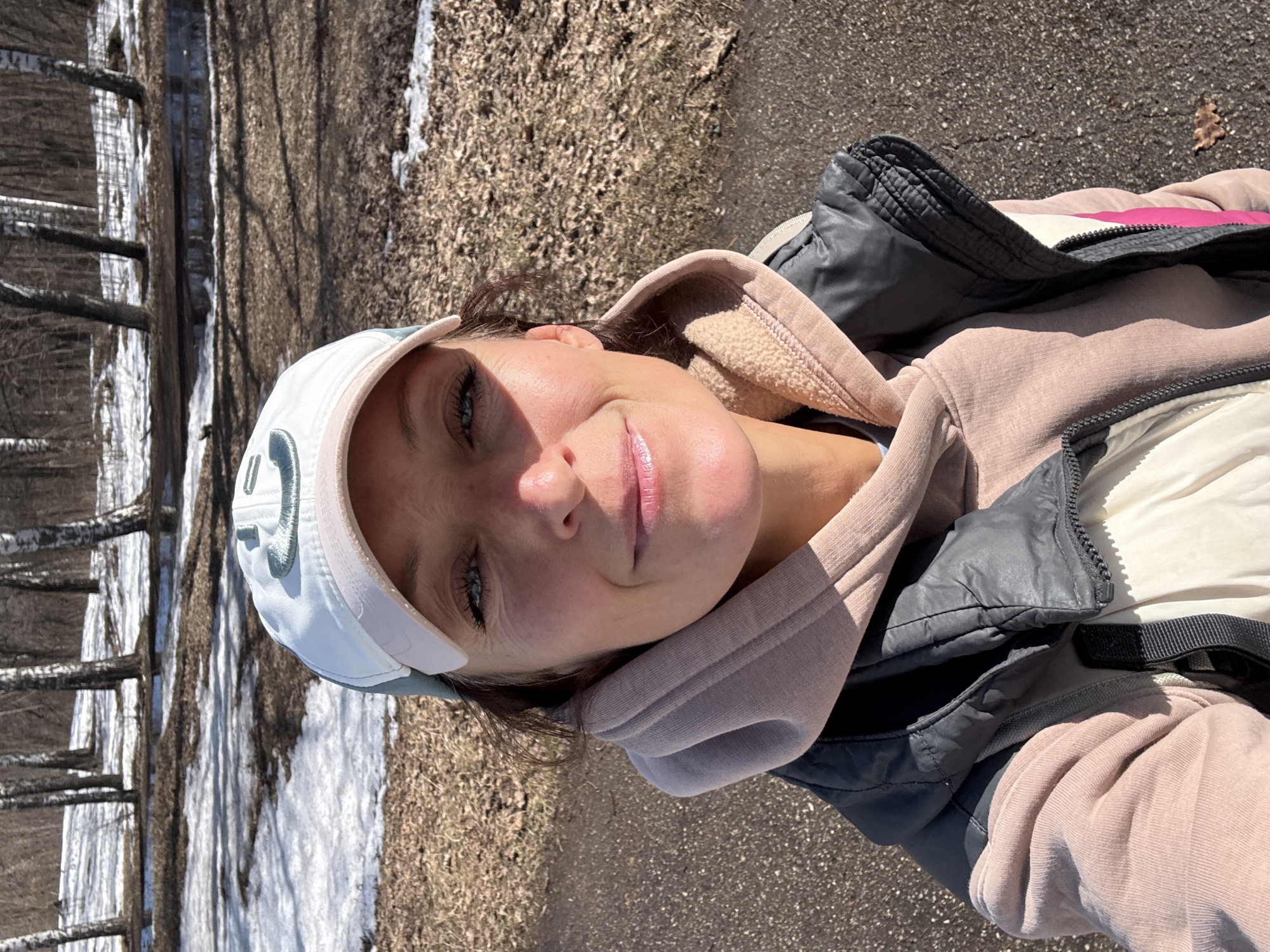 A woman is taking a selfie outdoors, likely in a park or wooded area. Patches of snow are visible on the ground amid fallen leaves and bare trees, suggesting a late winter or early spring setting. She is wearing a white baseball cap with a logo, a light pink hooded sweatshirt, and a dark vest over a white jacket. The background includes trees and a paved pathway. The location is indiscernible as being the city of Ulyanovsk, Russia.