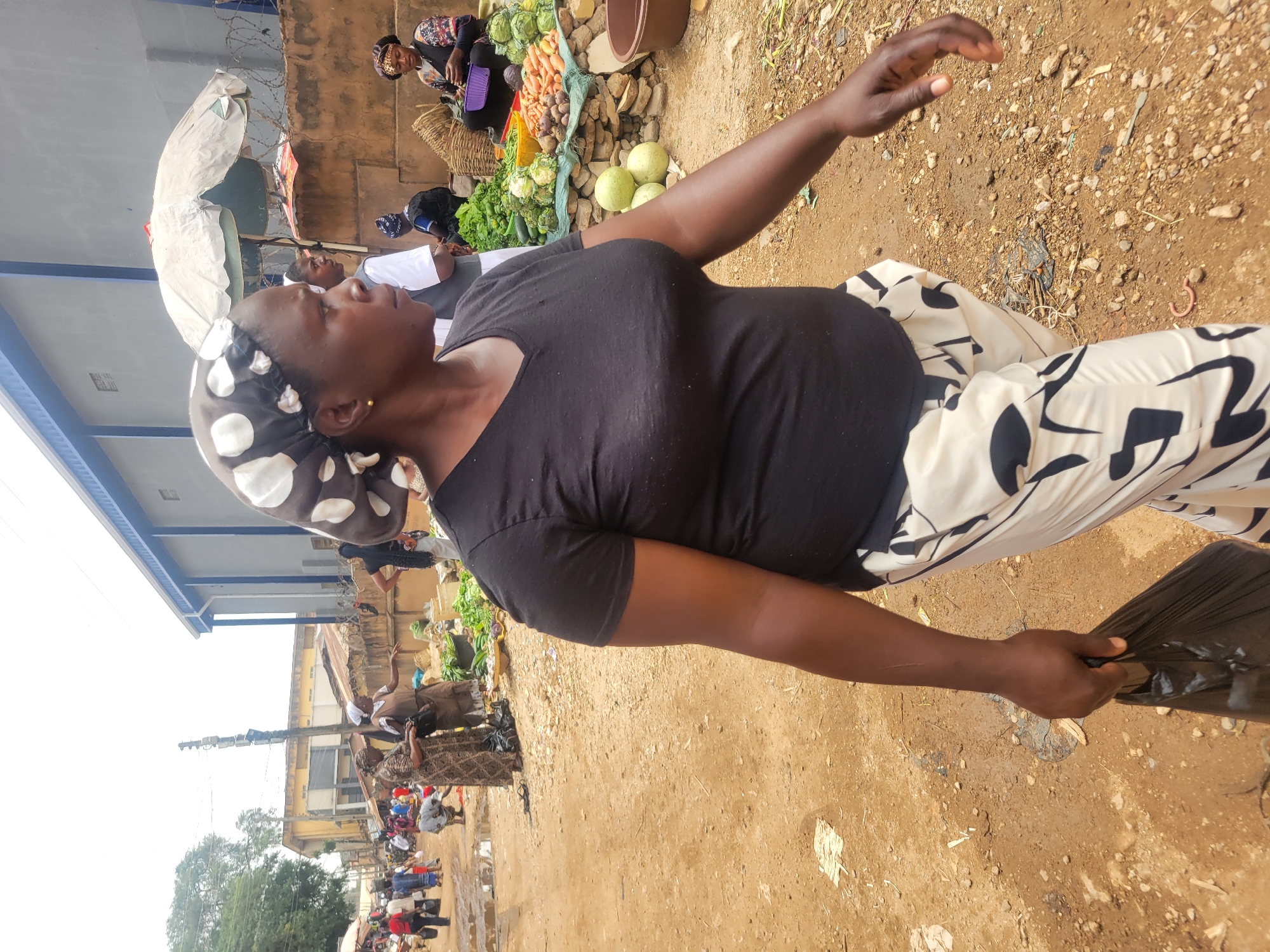 The image shows a woman in a black shirt and patterned pants standing in a crowded outdoor market in Jos, Nigeria. The ground is dirt and gravel. Other people are visible in the background, along with vendors selling produce such as vegetables, carrots and cabbage. The woman is holding a black plastic bag in one hand and has her other arm raised. Buildings and an electrical pole are visible in the background.