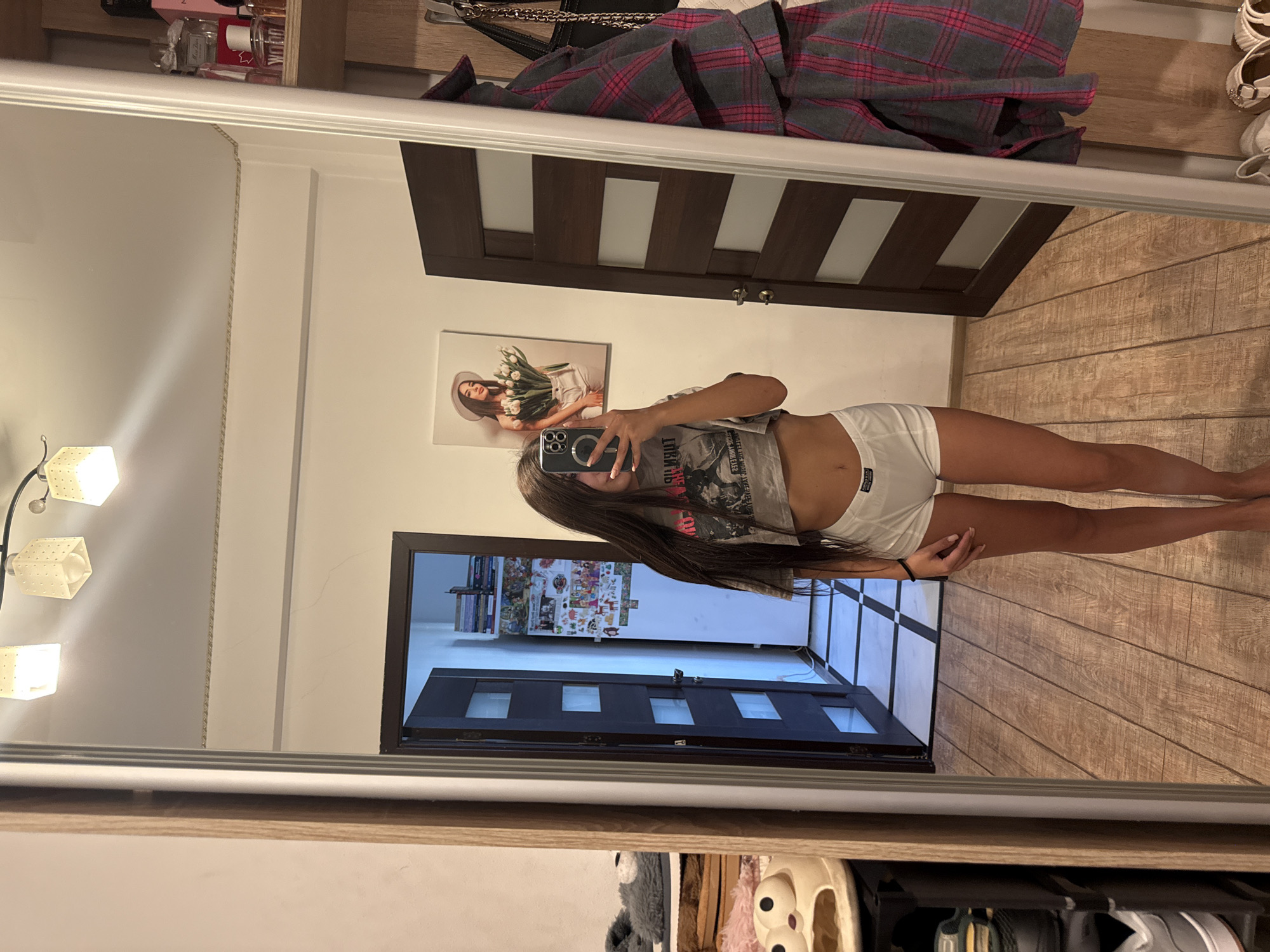 This is a medium shot of a young woman, seen from the waist down, taking a selfie in a mirror. She is wearing white shorts and a gray t-shirt. The setting appears to be an indoor space, possibly a bedroom or dressing area, with a wooden floor and a large mirror. In the reflection of the mirror, a closed wooden door with glass panels is visible, along with some artwork on the wall. The overall lighting suggests it might be daytime, but the specific time or weather is not discernible. There is no discernible text in the image.