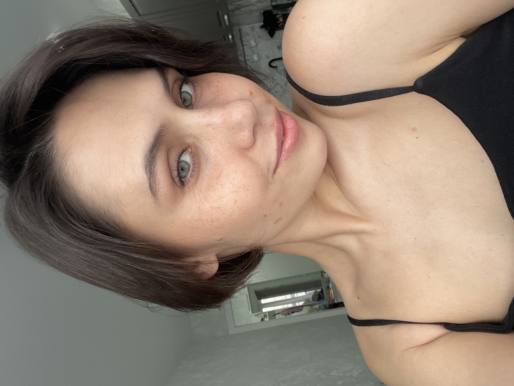 A close-up selfie captures a young woman positioned at a dynamic, tilted angle, looking directly into the camera with a soft, engaged expression and a slight smile. She has short, dark brown hair that frames her face, and striking light greenish-gray eyes. Her fair skin features numerous freckles across her nose and cheeks. She is wearing a simple black strap top, with her bare shoulder and upper chest visible, highlighting her collarbone. The lighting is bright and natural, suggesting the photograph was taken during the daytime indoors. The background is softly blurred but appears to be a light-colored room, possibly with a mirror or window reflecting light in the distance. No visible text is present in the image. This media file originates from Izhevsk, Russia.