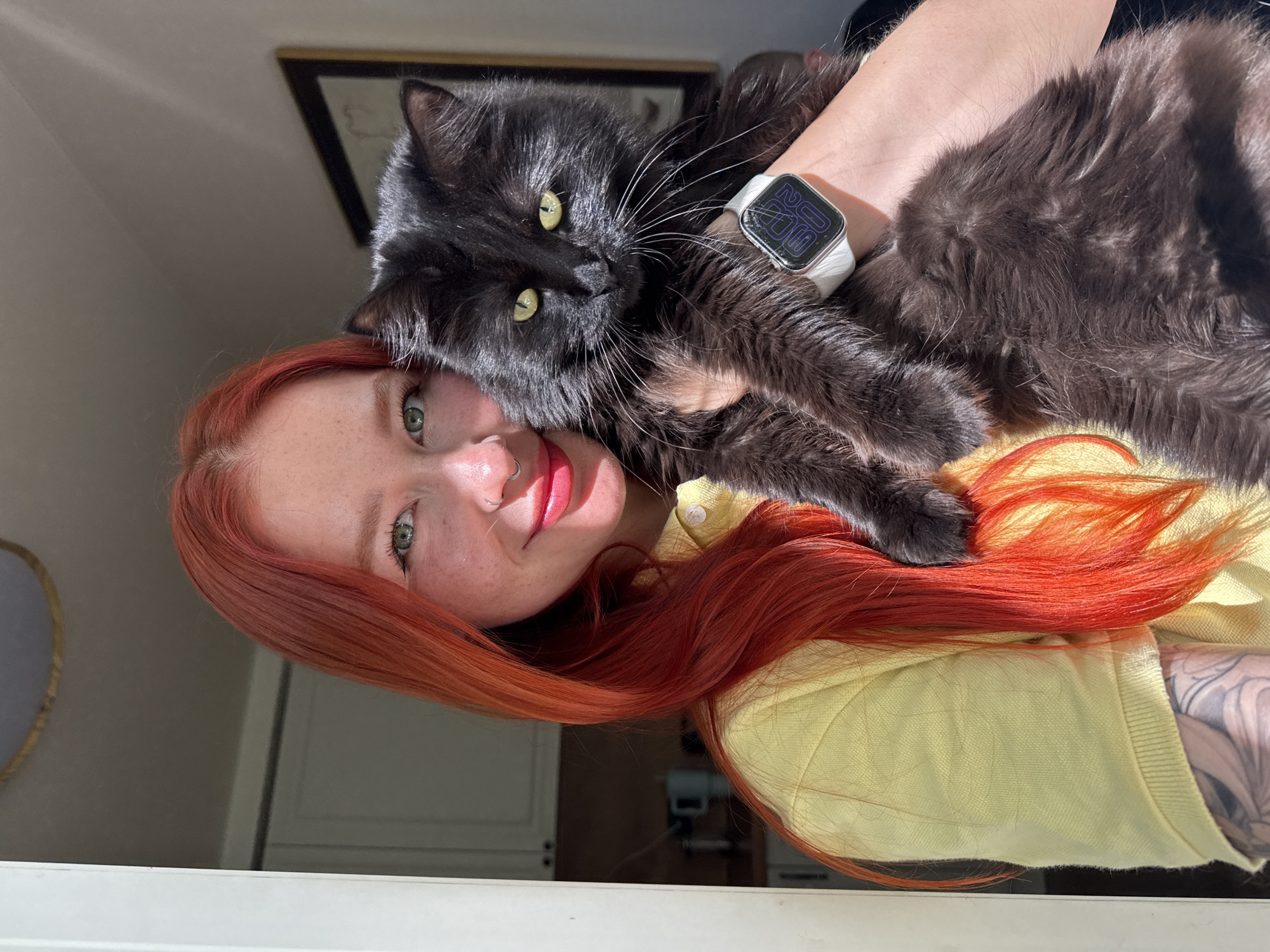 A woman with red hair is holding a fluffy black cat. The cat is resting on the woman's chest and shoulder, with its paws near her face. The woman is wearing a light yellow top. On her wrist is a smartwatch with a white band. There is a window in the foreground, and a picture frame on the wall in the background. The photograph appears to be taken indoors. I am unable to comment on the location in Rostov-on-Don, Russia, as there are no details confirming this in the image.