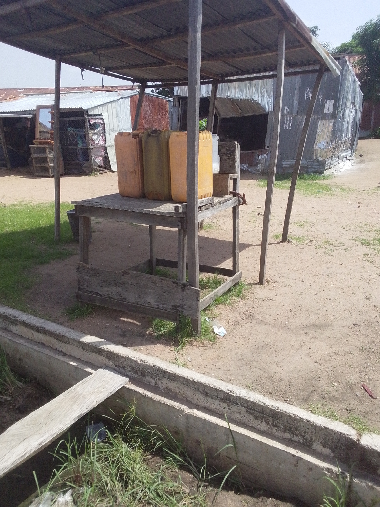 The image depicts an outdoor scene in Jalingo, Nigeria. In the foreground, a concrete structure, possibly a drainage channel, is visible with some vegetation growing alongside it and a wooden plank extending over it. In the midground, a rustic wooden table stands under a corrugated metal roof supported by wooden posts. On the table are three large plastic jerrycans, one orange and two yellow. The ground around the table is sandy with patches of grass. In the background, a building constructed with corrugated metal sheeting is seen, appearing somewhat dilapidated. The overall environment suggests a rural or semi-urban setting, possibly a market or storage area. There are no people visible in the image.