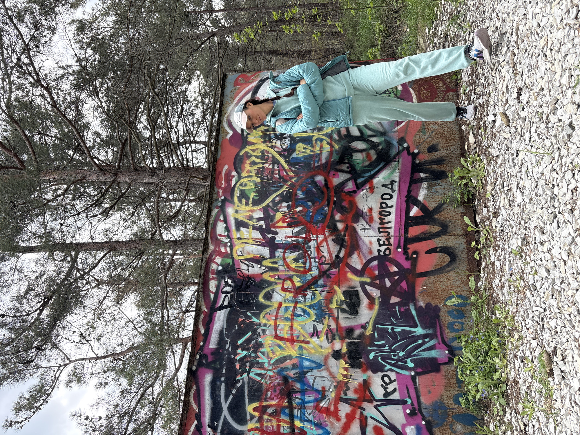 The image features a young person standing in front of a heavily graffitied wall in an outdoor setting, likely posing for a photograph.
The main subject is a young person, dressed in a comfortable, monochromatic light blue or mint green athletic set including sweatpants and a matching hooded top. They wear a light blue puffer jacket over this outfit, a light-colored baseball cap, and white sneakers with blue accents. A small dark bag is slung over their shoulder. The person has their arms crossed and looks towards the camera with a neutral expression.
The setting consists of a large, rusty wall, which serves as a vibrant backdrop, entirely covered in colorful graffiti. The graffiti includes a diverse mix of tags, symbols, and abstract designs in red, yellow, black, pink, blue, green, and white. A prominent red pentagram-like symbol is visible. Cyrillic text is present, most clearly "БЕЛГОРОД" (Belgorod) written vertically in black towards the right side of the wall, with "ГОРОД" forming part of it. Below this, the letters "КР" are also visible.
The ground directly in front of the wall is covered with light grey gravel or crushed stones, with some green weeds and small plants growing along the base. Behind the wall, a dense arrangement of pine trees with their branches and needles are visible against a bright, overcast sky, indicating it is daytime. The scene suggests an urban or semi-urban outdoor environment, possibly a recreational area or an abandoned structure, located in Gelendzhik, Russia.