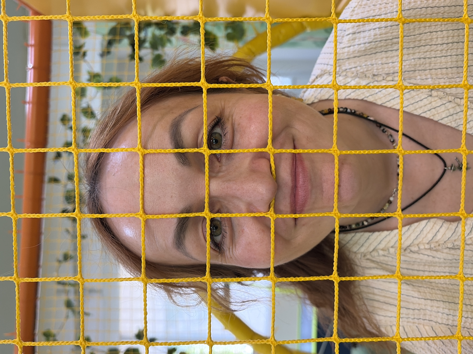 A woman with red hair is seen through a yellow net. The woman has green eyes and is wearing a striped shirt and a beaded necklace. The background appears to be an indoor playground with green foliage and orange poles. The location is the city of Chaikovsky, Russia.