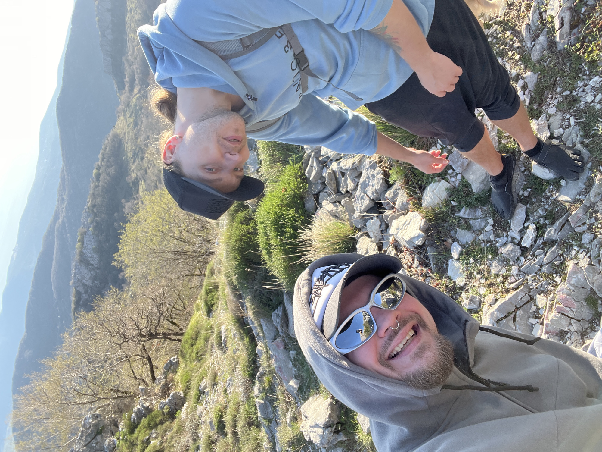 The image shows a selfie of two people on a rocky, mountainous terrain. In the foreground, a person wears a grey hooded sweatshirt, sunglasses, and a baseball cap. This person is smiling. Behind, another person, wearing a blue hooded sweatshirt, black shorts, and black toe shoes, is also visible. The landscape includes rocks, grass, and trees. In the background, there are mountains, suggesting a high altitude location. The image is potentially taken near Dolovi, Montenegro, based on the mountainous terrain.