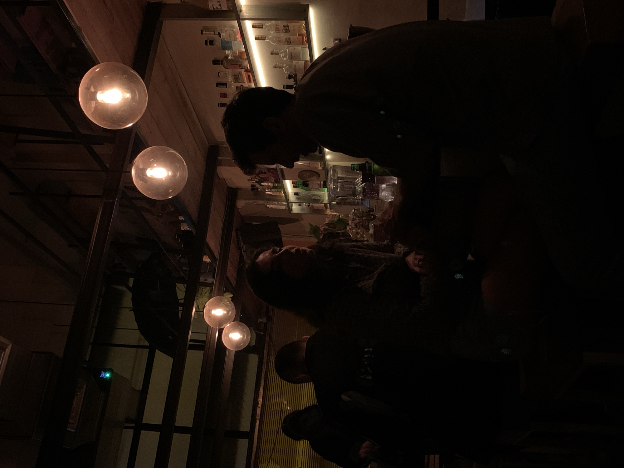 The image shows a dimly lit interior of a bar in Tel-Aviv, Israel. The bar appears to be well-stocked with a variety of liquor bottles. Three people are present at the bar. A bartender is shown bent over the counter, attending to a female patron. A third person stands adjacent to the female patron. Three spherical lights are suspended from the ceiling, illuminating the area. A reflection of the bar appears in a mirror.