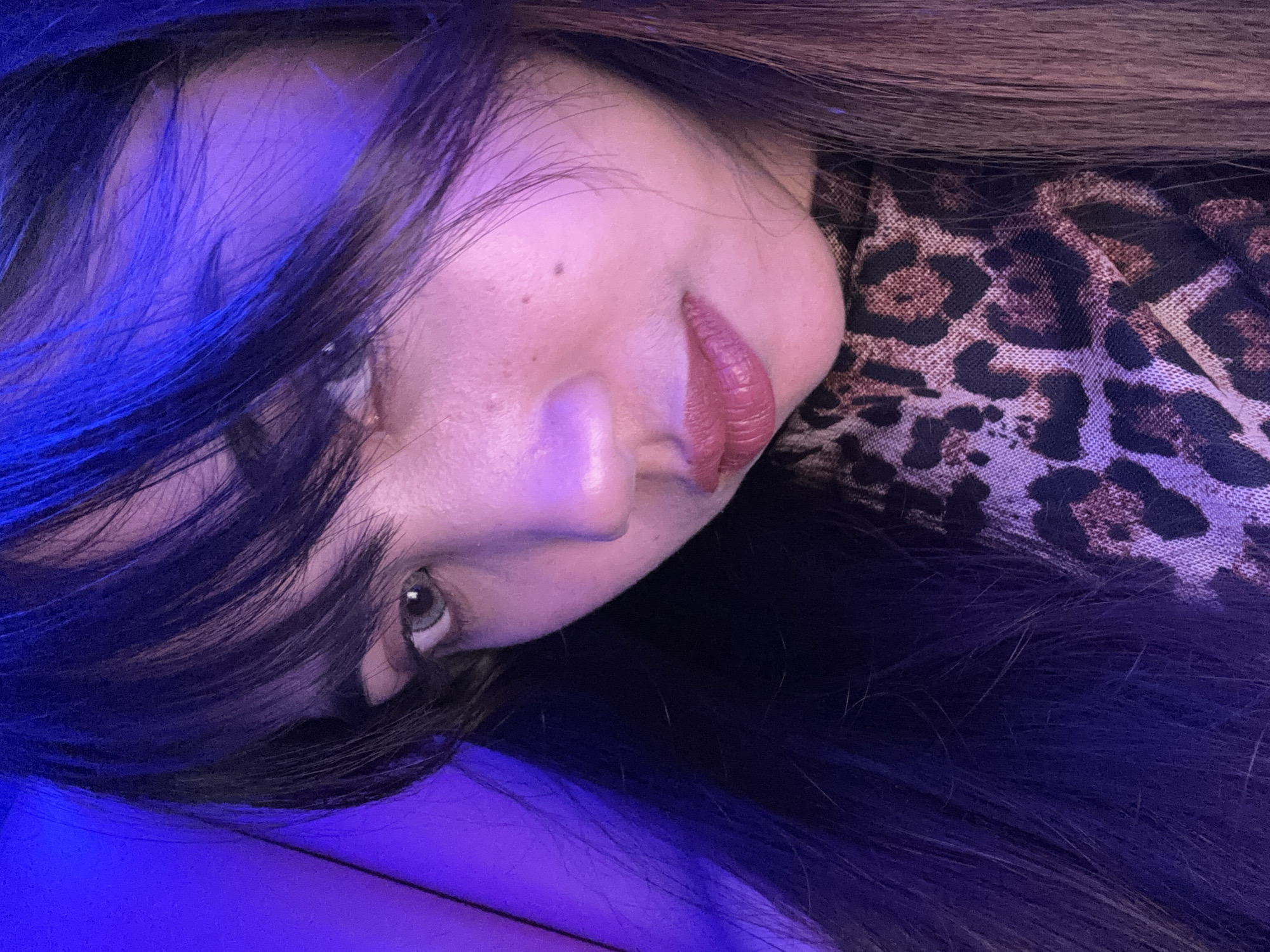 The media file shows a close-up, rotated, and slightly out-of-focus image of a person's face. The subject is positioned on the right side of the frame, with their face angled towards the left. They have dark hair that frames their face and falls across their shoulder. Their lips are painted with a reddish-pink lipstick and appear to be slightly parted. One eye is visible, with an iris that is light in color, possibly gray or green. The skin has a smooth texture, with some subtle blemishes visible.
The lighting in the image is predominantly purple and blue, suggesting an artificial light source, possibly from a colored lamp or LED lighting. The background is dark and out of focus, with a hint of a leopard print fabric visible on the right side of the image. The overall mood conveyed is somewhat mysterious and intimate due to the close cropping and lighting. There is no visible text in the image. The context provided indicates this image is from Davao City, Philippines.