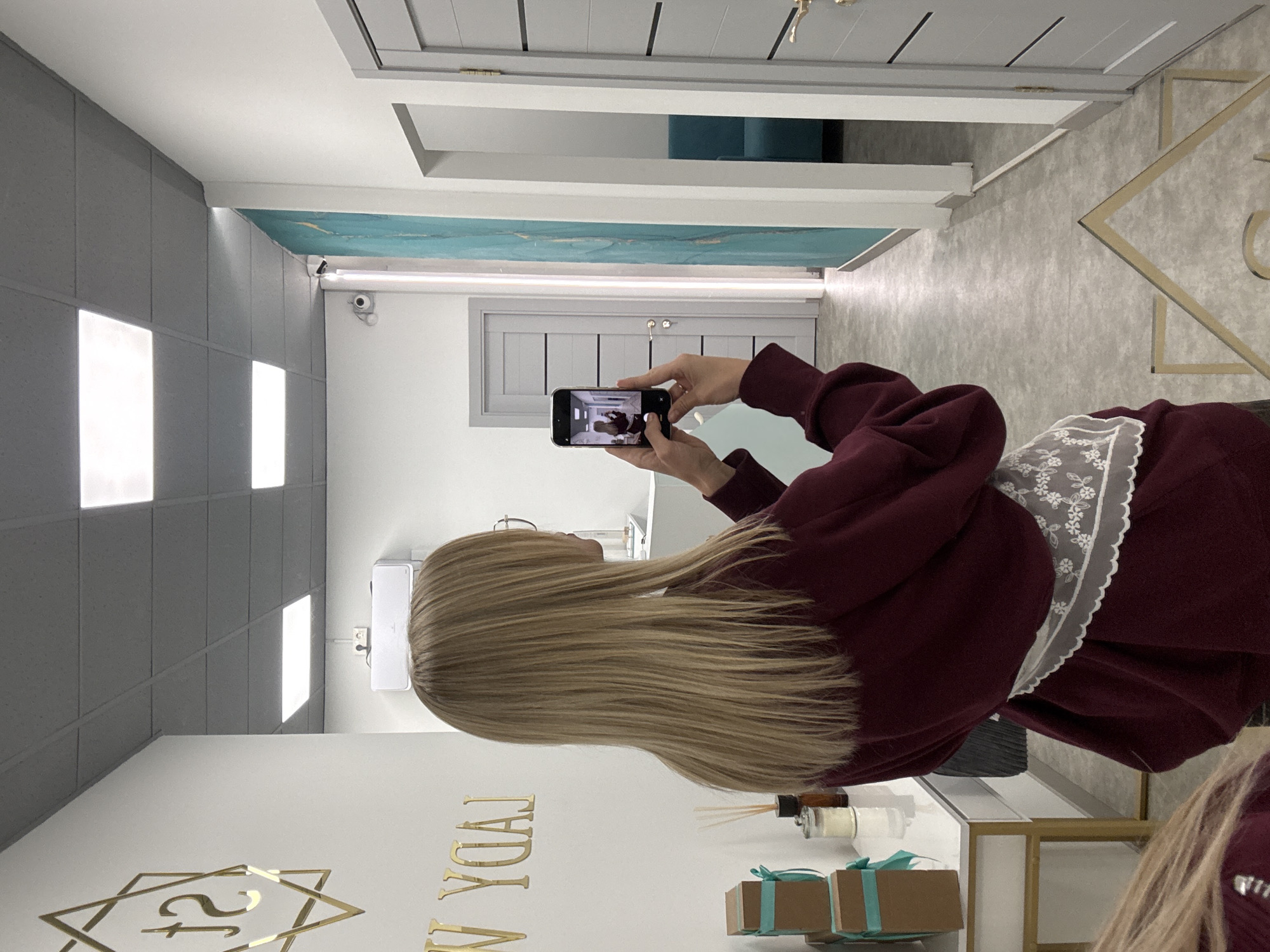 The image shows the interior of a commercial space. A woman is holding a smartphone, presumably taking a photo of the room. The room has a modern aesthetic with a gray ceiling and white walls featuring a gold logo for "Lady W." Decorations include small gift boxes with ribbons and scent diffusers. There are gray doors, one of which appears open to reveal a turquoise sofa. Security cameras are visible near the ceiling. The photo was taken in Krasnoyarsk, Russia.