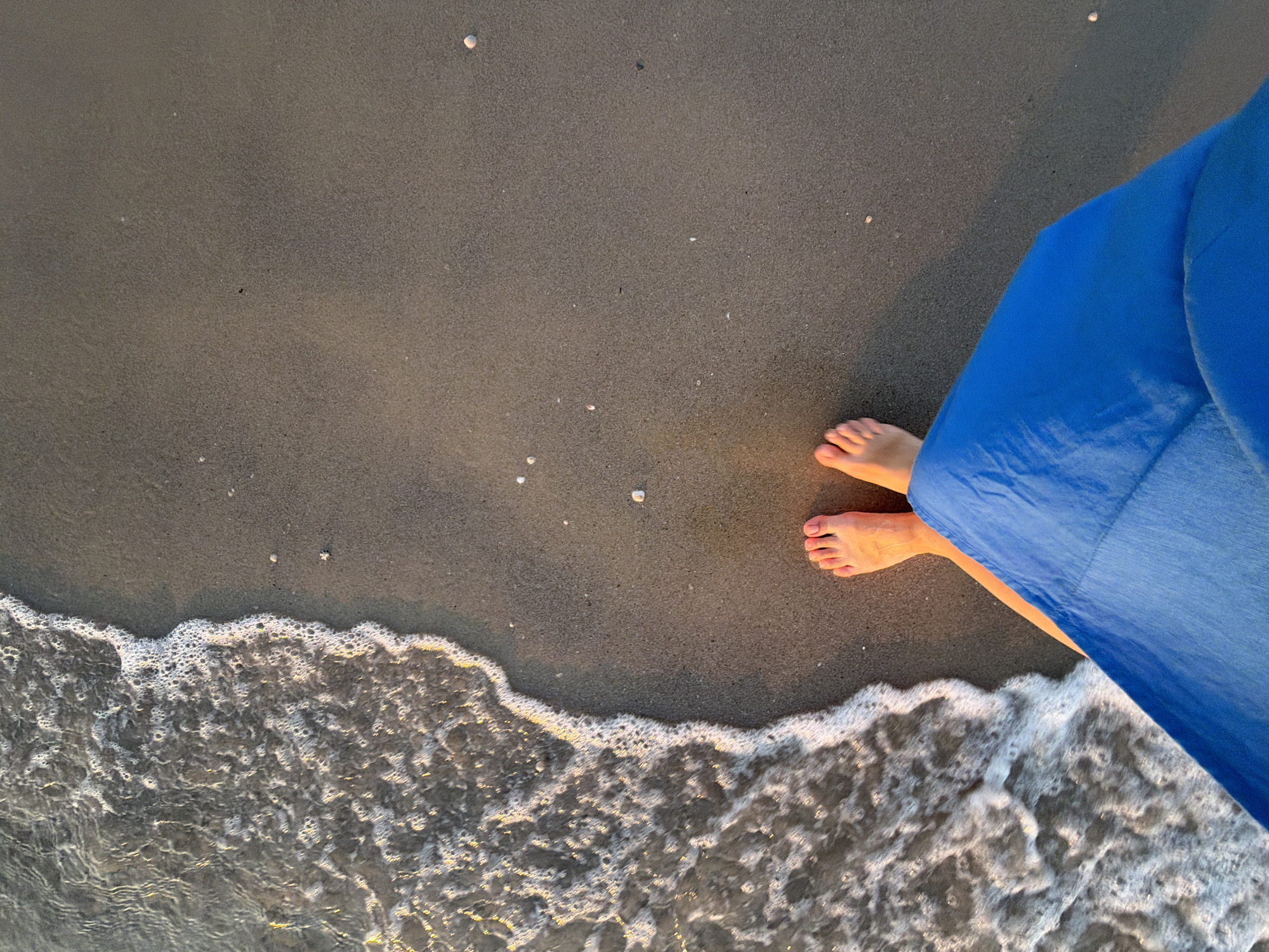 The image depicts a top-down view of a person's feet and a portion of a blue garment on a sandy beach in Bat Yam, Israel. Gentle waves with white foam are washing ashore on the left side of the frame, creating a dynamic waterline. The sand is a light brown, with scattered small shells visible. The person's bare feet, with visible toes and toenails, are positioned towards the bottom center of the image, stepping onto the wet sand at the edge of the receding wave. The blue garment, possibly a dress or skirt, occupies the lower right quadrant, obscuring the lower body of the person. The lighting suggests it is either early morning or late afternoon, casting soft shadows on the sand. The overall scene conveys a sense of peacefulness and the simple pleasure of being by the sea.
