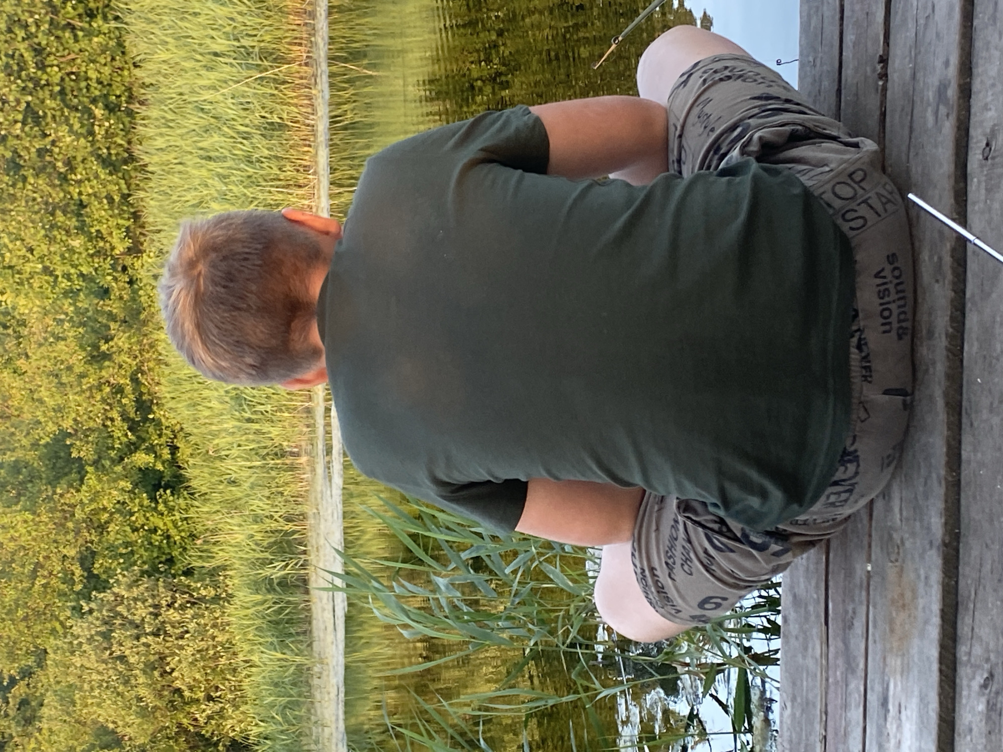 A man is sitting on a wooden dock, facing away from the camera. He is wearing a dark green t-shirt and camouflage shorts. The shorts have text printed on them, including "TOP STAR", "SOUNDS VISION", and "FASHION CHA". He appears to be fishing, with a fishing rod extending into the water. The setting is natural, with tall grasses and trees on the left side of the image, and calm water on the right. The water reflects the surrounding greenery and the sky, suggesting it's daytime with clear weather. The man's posture indicates relaxation and concentration on his activity. The wooden dock is weathered, with visible planks and nails. The overall scene conveys a sense of peaceful outdoor recreation.