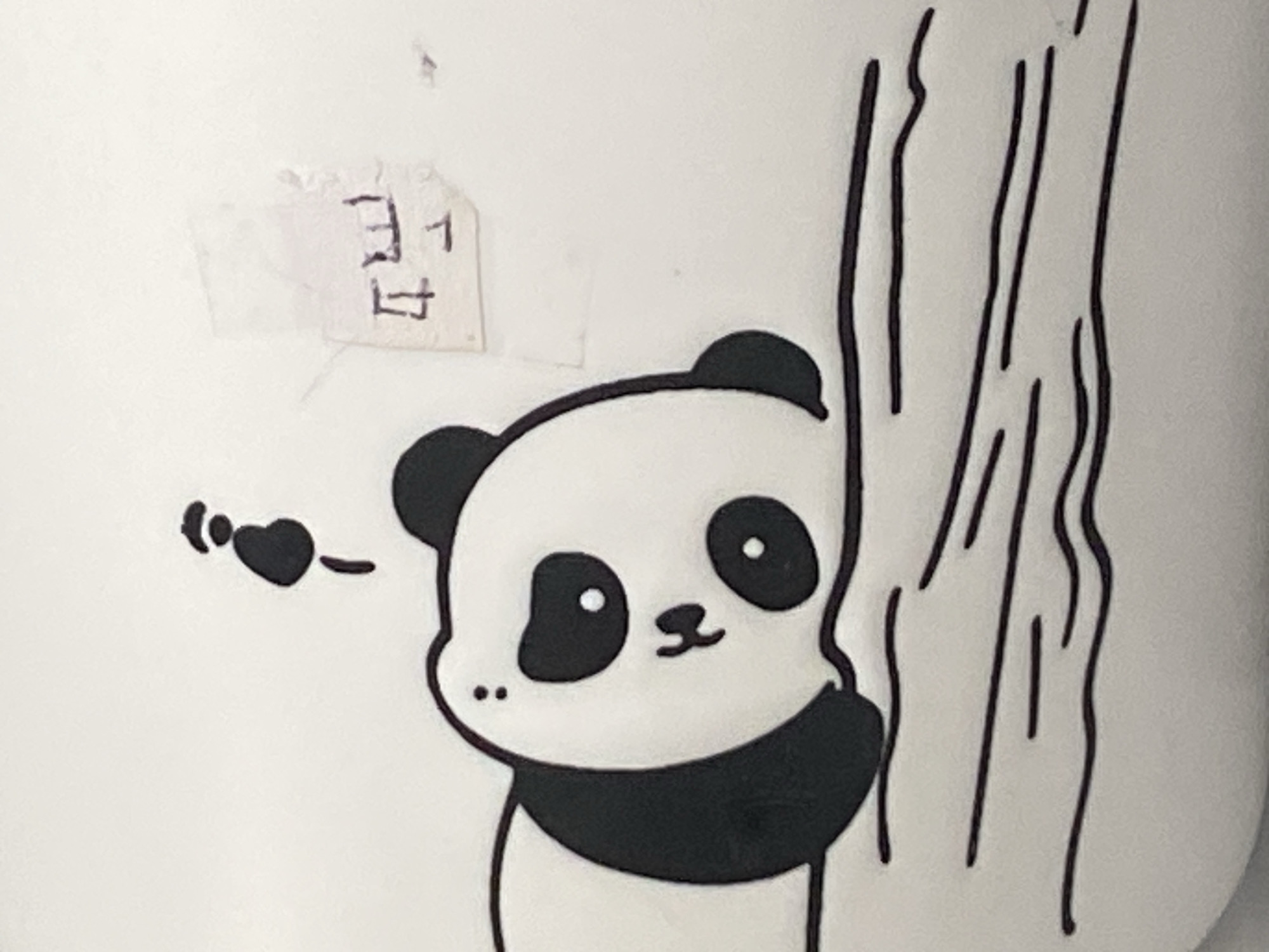The image displays a close-up view of a white ceramic mug or similar object adorned with a drawing of a panda peeking from behind a tree. The panda is depicted in a cartoonish style, with large black eye patches, a black scarf-like marking around its neck, and a small smile. A small black heart with a dashed line extending from it, possibly indicating a kiss or affection, is drawn to the left of the panda. The tree is represented by several vertical, wavy black lines. There is also a small piece of paper with some writing on it taped to the white surface in the upper left quadrant of the image. The writing appears to be in a foreign script and is partially obscured. The lighting is even, and the focus is sharp, highlighting the details of the drawing. The image does not provide any information about its location in the city of Rybnitsa, Moldova.