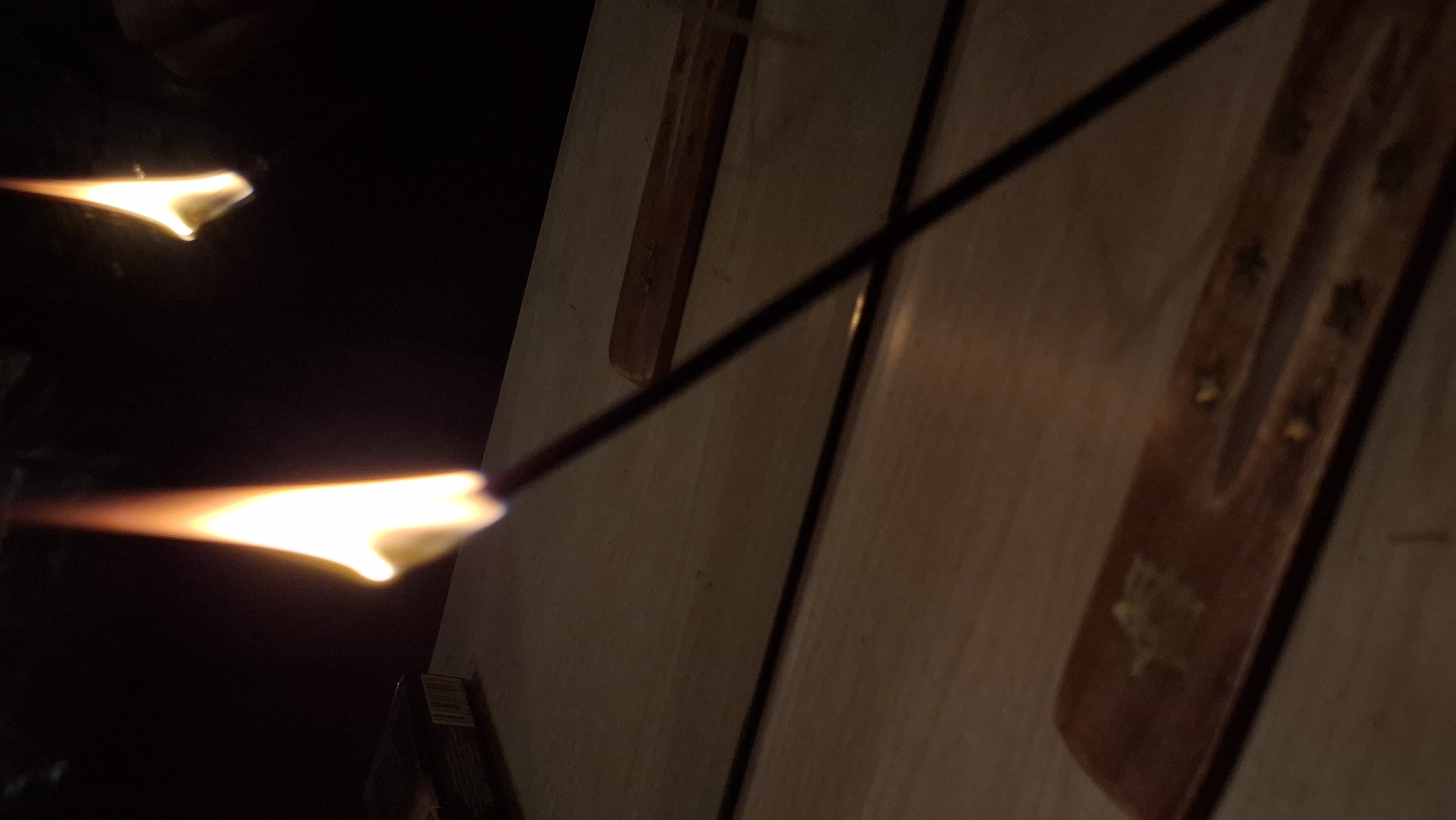 The image depicts a close-up, low-light view of an incense stick burning, with its flame prominently featured. The incense stick is held at an angle, extending diagonally across the frame from the bottom right towards the top left. The flame is bright orange and yellow, with wisps of smoke faintly visible. In the background, a dark surface is present, possibly a wall or a dark cloth. To the left of the flame, a portion of a black box with some white markings is visible, suggesting it could be a cigarette pack or a similar item. The foreground shows a wooden surface, likely a table or shelf, with a faint reflection of the flame. There is also a wooden incense holder with indentations designed to hold incense sticks, visible on the wooden surface. Another small, out-of-focus flame is reflected in what appears to be a mirror or reflective surface, positioned in the upper right background. The overall ambiance is dim and intimate, illuminated primarily by the burning incense. There are no discernible people or animals present in the image. The scene is indoors, possibly in a residential setting, and the location within Забалоцкі сельскі Савет, Belarus cannot be determined from the visual information provided.