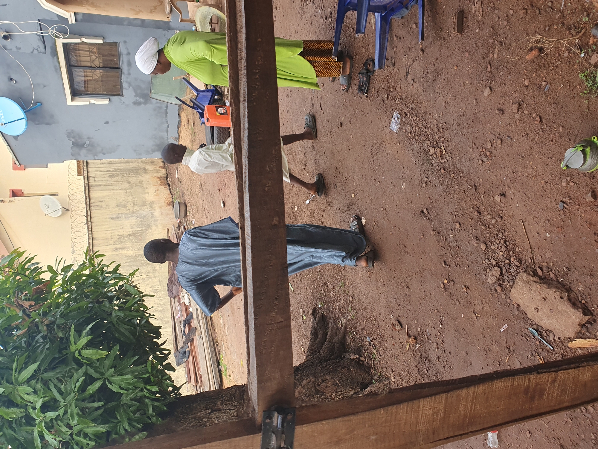 This media file captures an outdoor scene, likely an open yard or pathway between buildings, with a reddish-brown dirt ground. Three individuals are present. One person, on the right, wears a white head covering and a bright green long-sleeved top with a patterned skirt, and black sandals. Another individual, closer to the center, is dressed in a light-colored (possibly white or cream) top and dark trousers, wearing black sandals or slip-on shoes. The third person, on the left and partially obscured by a wooden beam, wears a blue or grey striped tunic and trousers, also with sandals.
The activity appears casual, with individuals standing or moving in the space. The person in green is positioned near blue plastic chairs and an orange jerry can. The person in the light top seems to be facing the person in blue/grey.
Notable details include a light-colored building in the background featuring a dark window and a blue satellite dish with the text "TSTV" visible on it. Another building with a fence topped with barbed wire is also visible. A large, leafy green tree is on the left. The foreground is dominated by prominent wooden beams or posts. Scattered on the dirt ground are various items such as small stones, debris, a plastic bag, a blue plastic spoon, and a plastic bottle with "AQUA" and "WATER" partially visible. The scene is illuminated by natural daylight. The overall setting, clothing, and architectural elements are consistent with Idah, Nigeria. Emotions are not discernible due to the distance and lack of clear facial expressions.