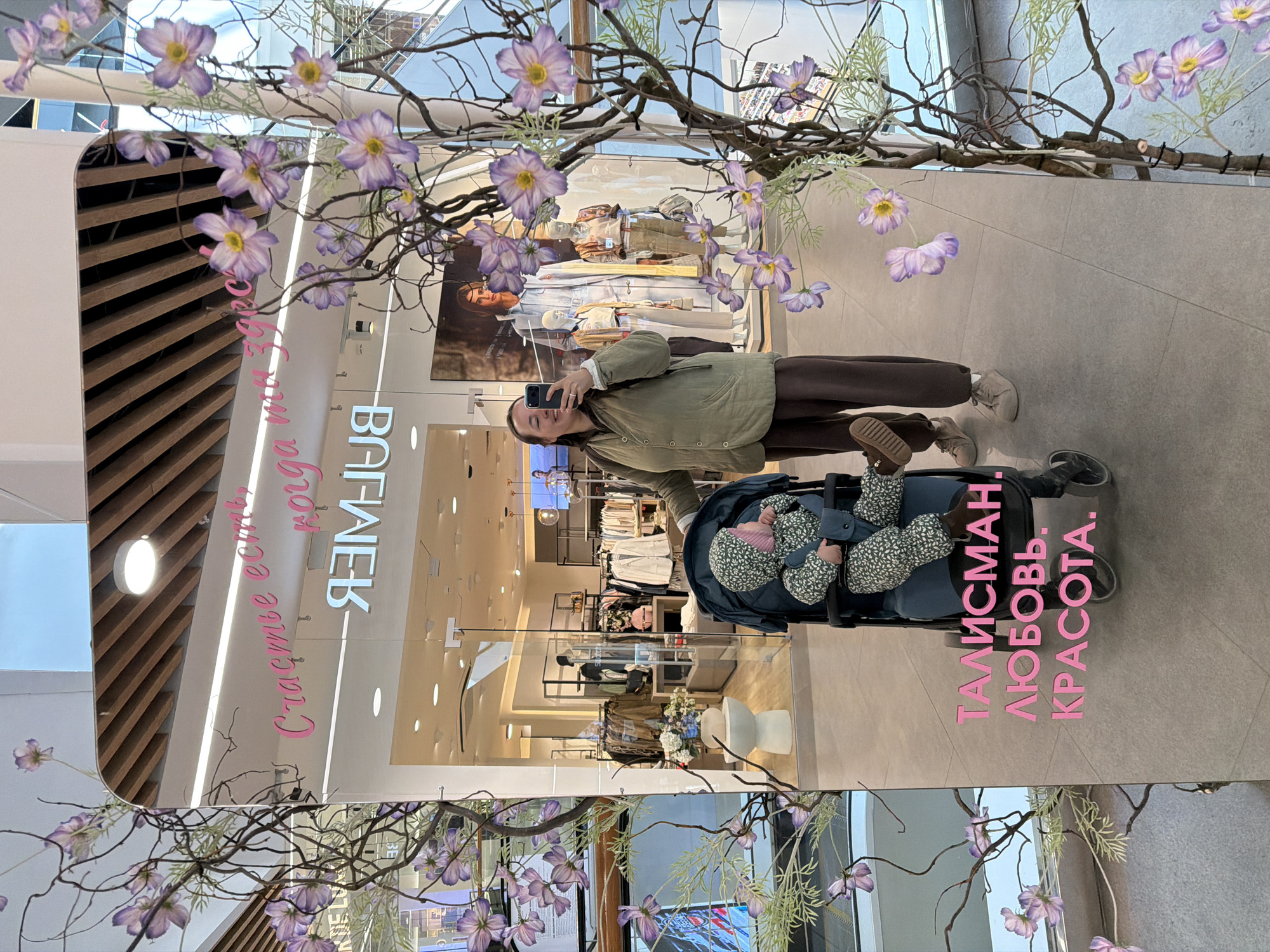 A woman wearing an olive-green jacket and brown trousers holds a mobile phone in her right hand, presumably taking a mirror selfie in a retail environment. In front of her is a stroller with a baby inside wearing patterned clothing. The store has clothing on display, mannequins, and floral arrangements. The mirror contains text in Russian, including "Счастье есть когда мы здесь" and "ВАГНЕВ" in reverse. On the ground near the stroller, there is additional Russian text that translates to "Talisman, Love, Beauty." The scene appears to be located in the city of Izhevsk, Russia, based on the store name.