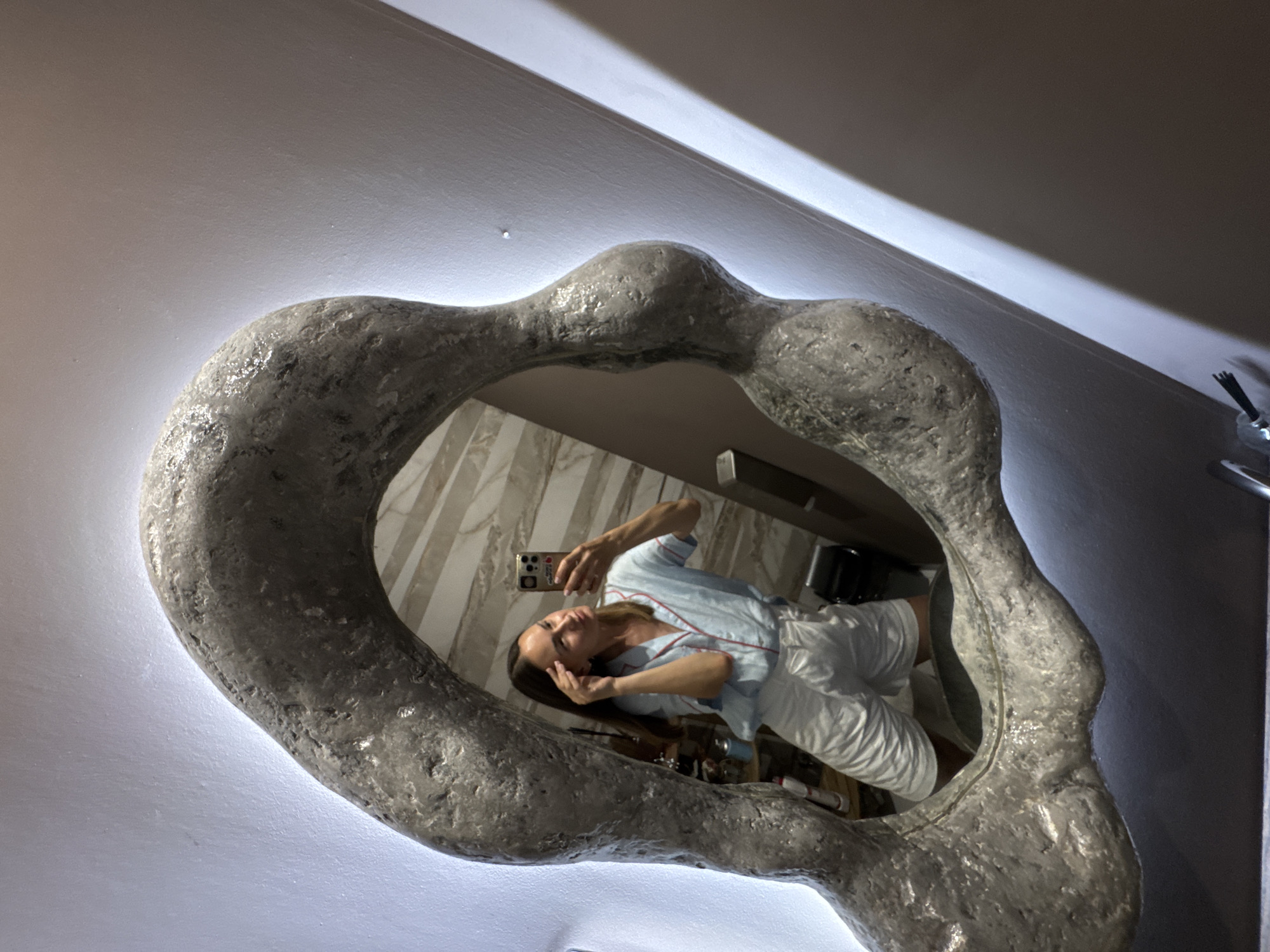 A woman is taking a selfie in a mirror in what appears to be a bathroom. She is wearing a light blue pajama set with red piping and white shorts. She is touching her hair and looking at her reflection. The mirror has an irregular, organic-shaped frame that appears to be made of stone or concrete and is illuminated from behind. The wall behind the mirror is white, and the overall lighting is dim. There are some objects visible in the reflection in the background, including what looks like a shelf with bottles. The location is specified as Sochi, Russia.
