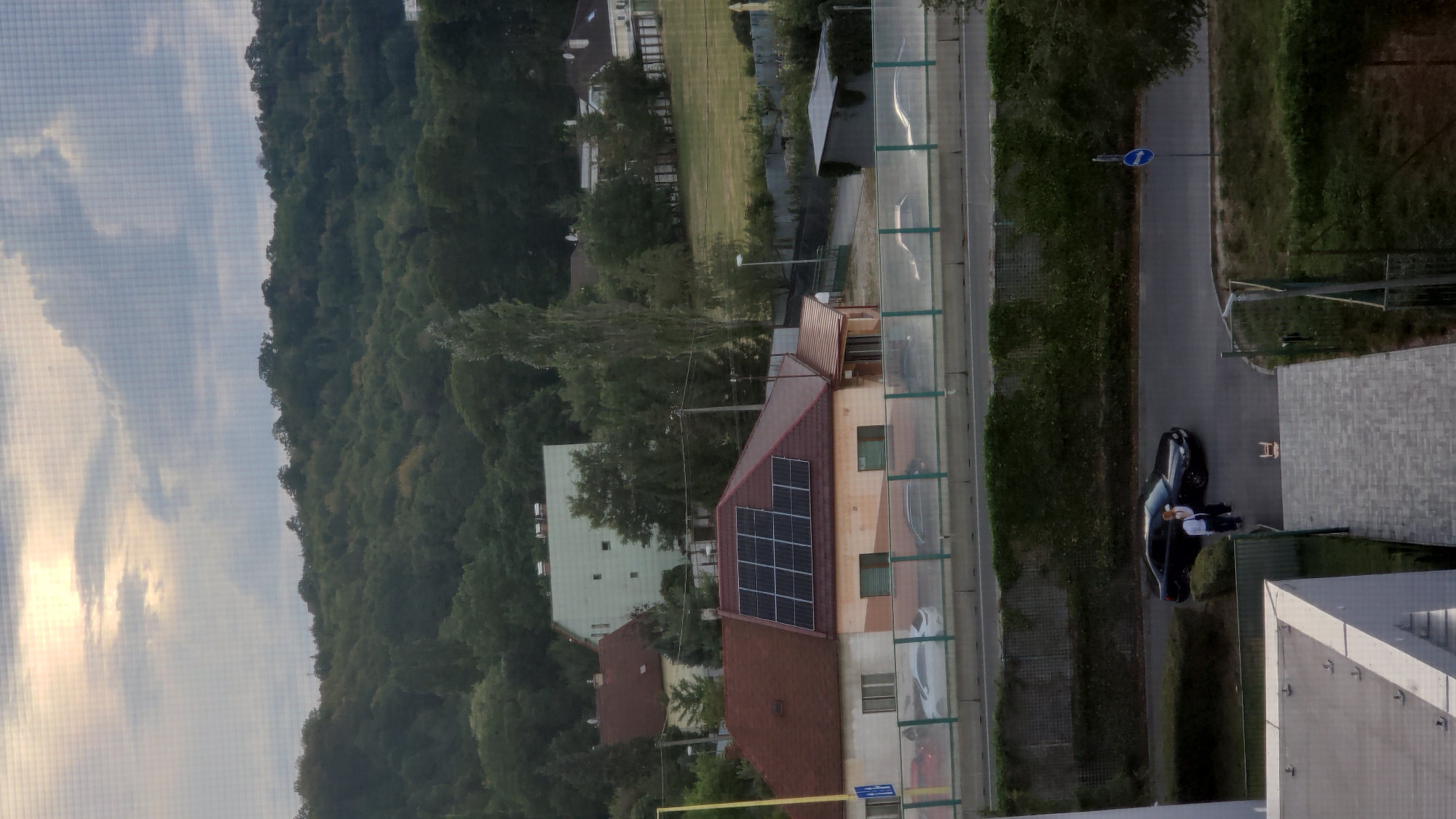 The image shows an overhead view of a suburban area with a dense forest in the background and a highway in the middle ground. On the left side of the highway, a house with solar panels on its roof is visible. Further down the highway, a black car is parked on the side of the road with two people standing next to it. A blue traffic sign with a white arrow pointing left is also visible near the car. The overall scene suggests a tranquil evening in a residential area with a natural landscape.