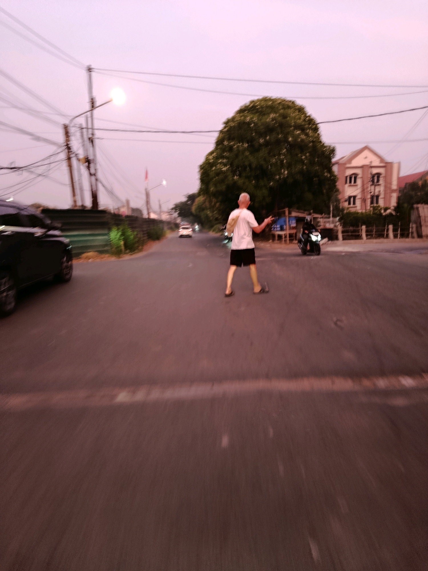 The image depicts a street scene at dusk in Bekasi, Indonesia. A blurry, middle-aged man with graying hair, wearing a white t-shirt and black shorts, is walking across the asphalt road. He has a bag slung over his left shoulder and is gesturing with his right hand. In the background, a person is riding a motorcycle with its headlights on, heading towards the man. To the left, a dark-colored SUV is parked. Further in the background, a large, lush green tree stands prominently, with a building featuring multiple windows and a reddish-brown roof visible behind it. Power lines and a street lamp with a bright light are also present, contributing to the twilight atmosphere. The road appears wet in places, and there are some construction barriers and fences visible along the side of the road. The overall impression is one of a typical urban evening in Bekasi, characterized by traffic, pedestrian activity, and the transition from day to night.