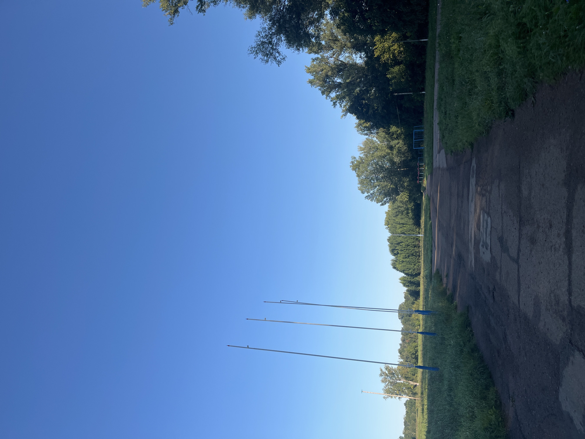 The image depicts an outdoor scene with a clear, bright blue sky dominating the upper half. In the middle ground, a dirt path leads away from the viewer into a grassy field. Several tall, slender poles, possibly for a sports field or lighting, are visible on the left side of the path, some with blue accents at their bases. Beyond the field, a line of trees forms a dense border. On the right side of the image, more trees create a darker, more shaded area. The ground in the foreground is a rough, patched surface. In the distance, on the left, a solitary lighter-colored pole is visible, and further back, a row of green trees stretches across the horizon. The overall impression is a peaceful, open space with elements of athletic or recreational infrastructure. No people are present in the image. The location is the Komsomolsky Park in Ufa, Russia, characterized by its open fields, mature trees, and recreational amenities.