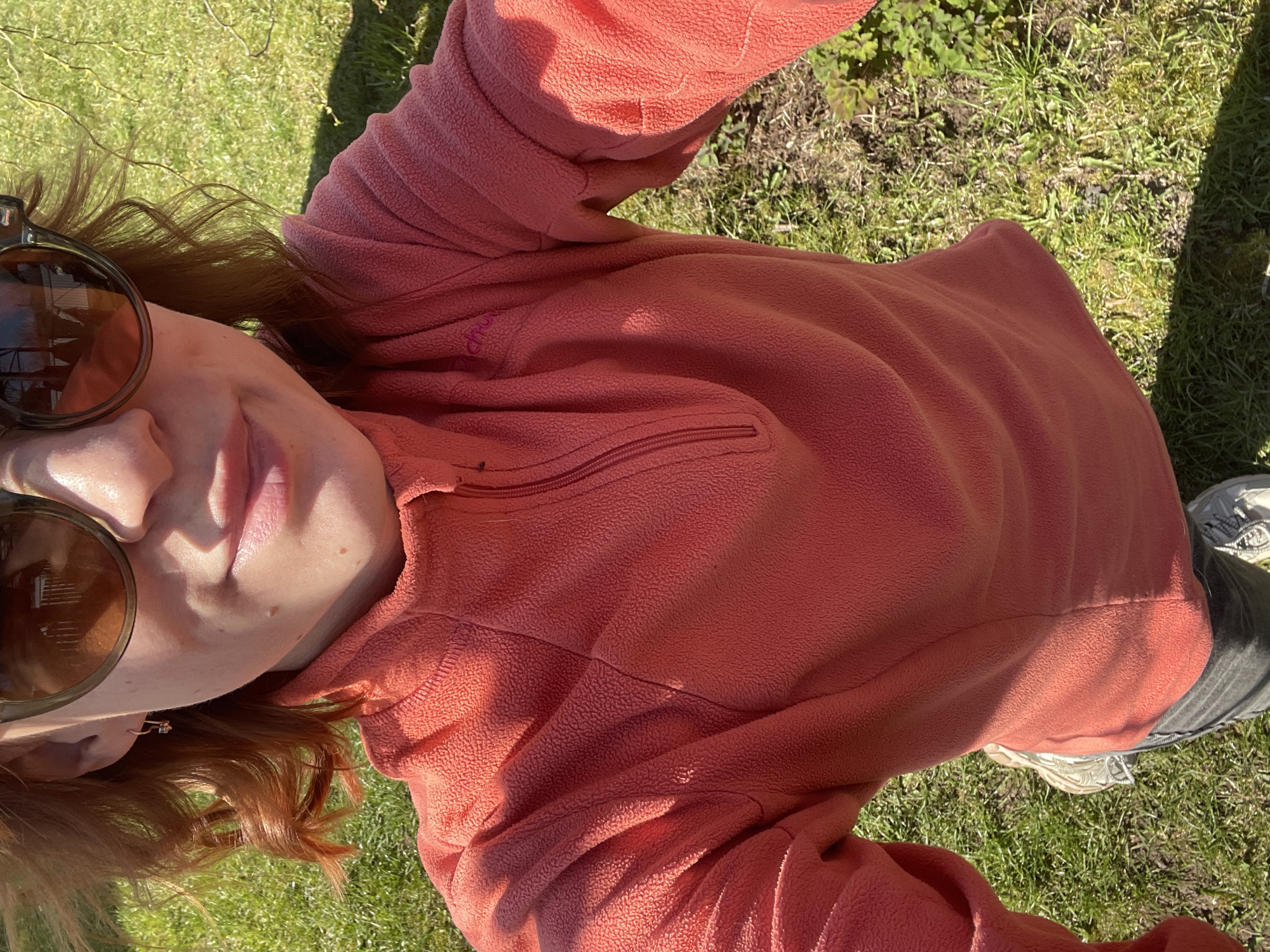 A person wearing a salmon-colored fleece top and sunglasses is standing on grass. The person has reddish-brown hair and a fair complexion. The background consists of a lawn area with both grass and bare patches of dirt. The lighting suggests a sunny day, with shadows cast on the grass and the person's clothing. The location within the city of Chaikovsky, Russia, cannot be determined from the image.
