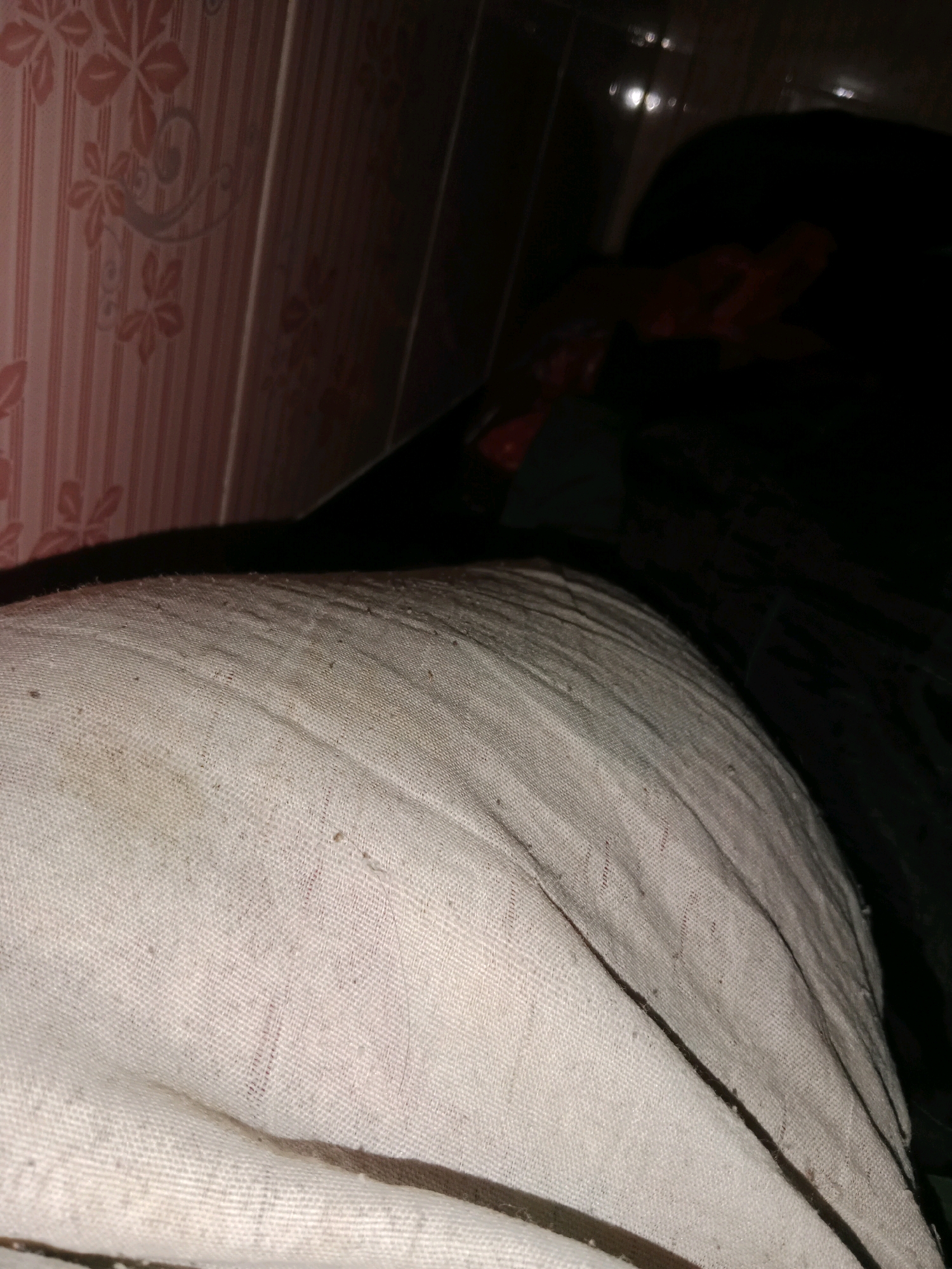 The image shows a close-up view of a person's leg, bandaged and covered with a white cloth. The bandage appears to be made of a textured material, possibly gauze or a similar fabric, with some dirt and debris visible on its surface. The leg is positioned diagonally across the frame, with the bandaged area taking up the majority of the foreground. In the background, a wall with a pink, striped pattern and floral motifs can be seen. To the right of the wall, the scene transitions to a dark area, possibly a room or a darker section of the environment. There are some blurred bright spots in the dark background, which could be reflections or light sources. The overall impression is that of a medical situation in a domestic or indoor setting. Without additional context, it is impossible to determine the specific location within Ringinrejo, Indonesia, beyond it being an indoor scene.
