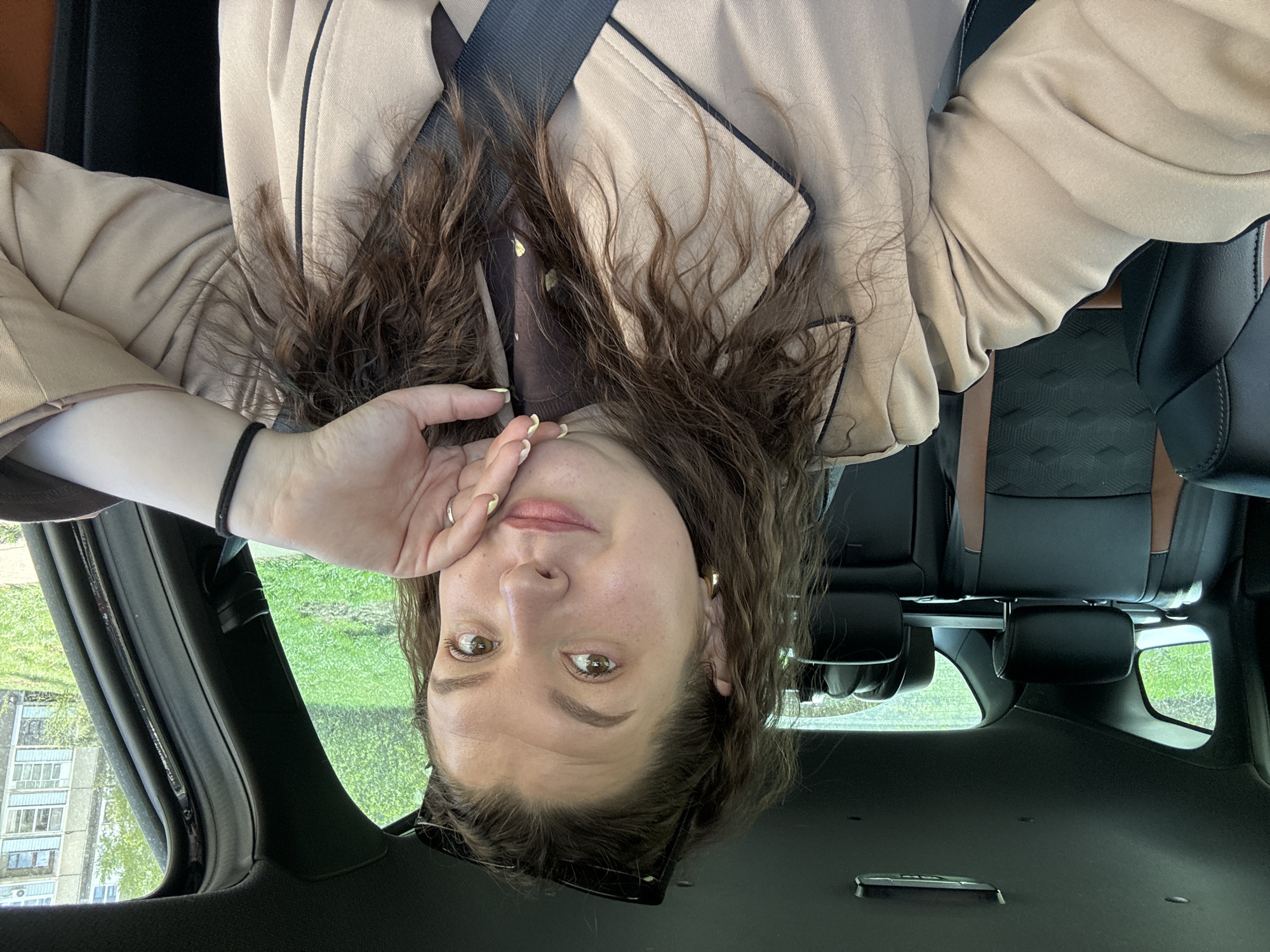 A woman is in a car, likely a passenger, wearing a tan-colored blazer with black trim, and a brown top underneath. She is wearing a seatbelt. Her dark curly hair is partially obscuring her face. Her left hand is near her face and she is looking at the camera. A black bracelet is on her wrist. Outside the car window is a green lawn and a multi-story residential building. The image is taken in a residential district that is consistent with the general architectural style of Марковский, Russia. The interior of the car features black leather seats with brown accents.