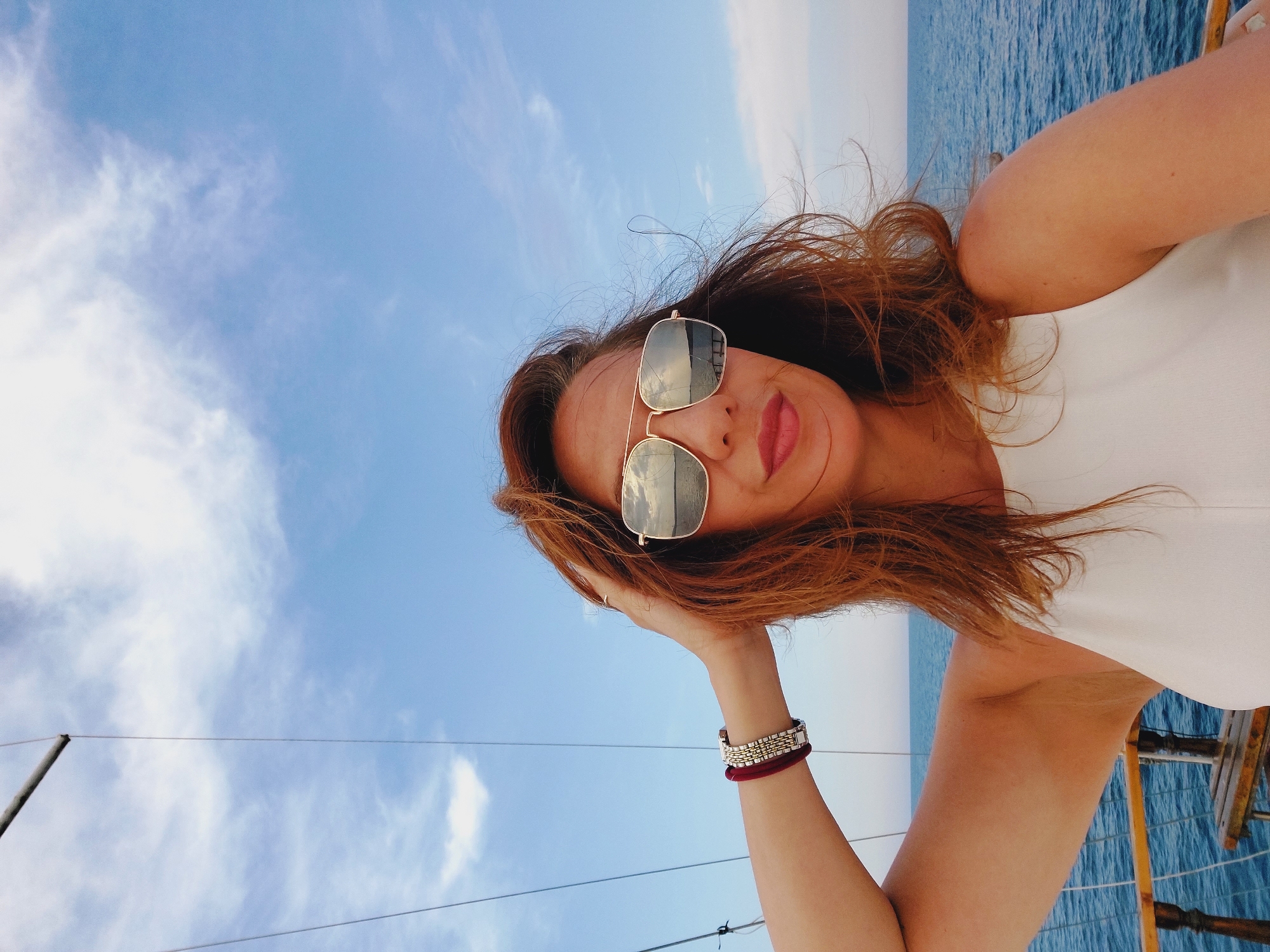 A woman with reddish-brown hair and sunglasses is taking a selfie while on a boat. She is wearing a white top and a bracelet on her left wrist. Her hand is raised, touching her hair. The background shows a blue sky with scattered clouds and the sea. The location is on a boat in the sea near Sozopol, Bulgaria.