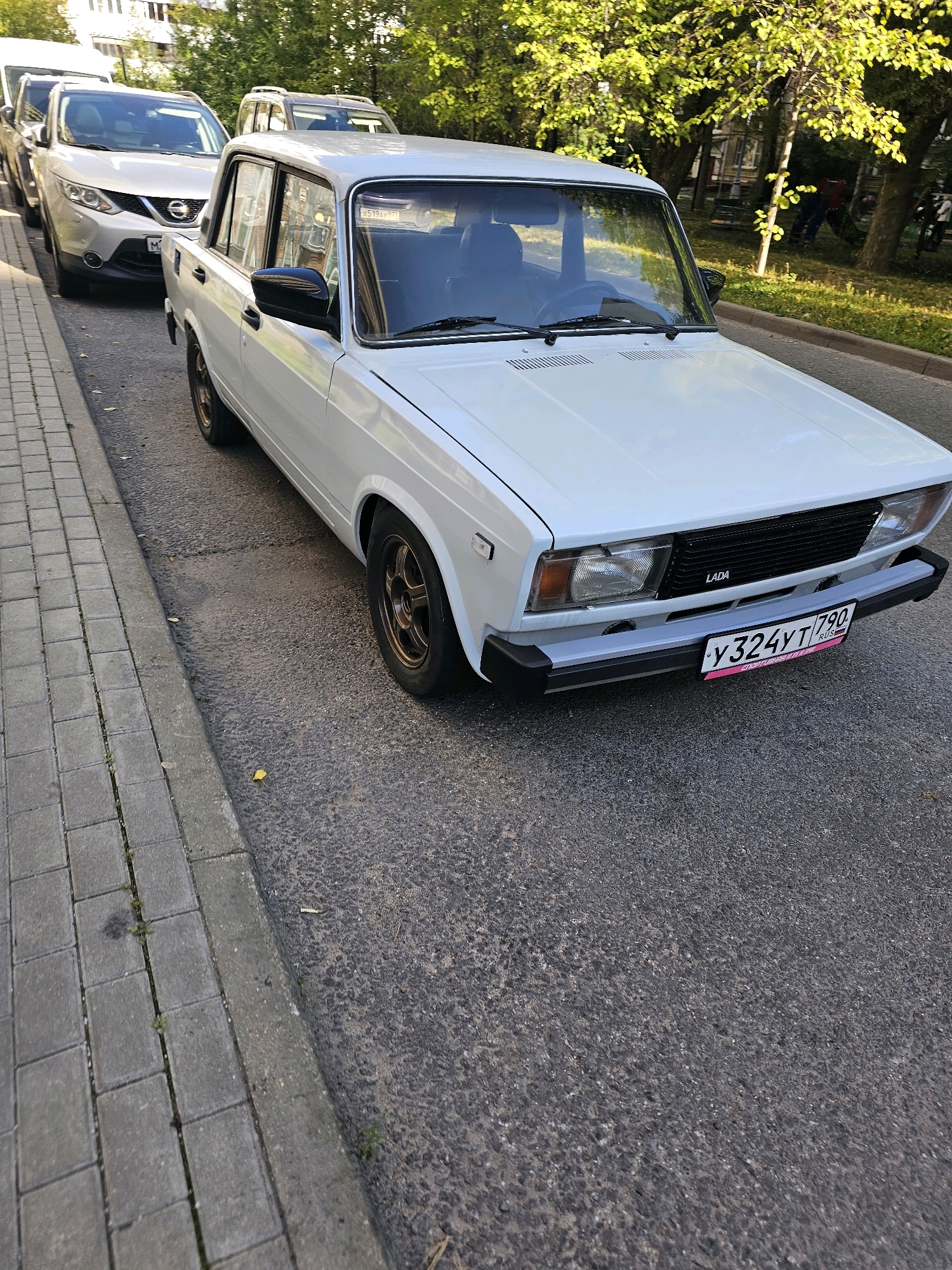 In Khimki, Russia, on a sunny day, a white Lada car is parked on the side of a paved street, adjacent to a sidewalk with gray square pavers. The car is in the foreground, angled towards the viewer, with its front bumper and grille clearly visible. A license plate with Cyrillic characters and the numbers "У324УТ 790" is attached to the front. Behind the Lada, other cars are visible, including a silver SUV. Trees with green foliage line the street, casting dappled shadows. In the background, there are residential buildings and a few people can be seen in the distance. The scene appears to be a typical urban streetscape.