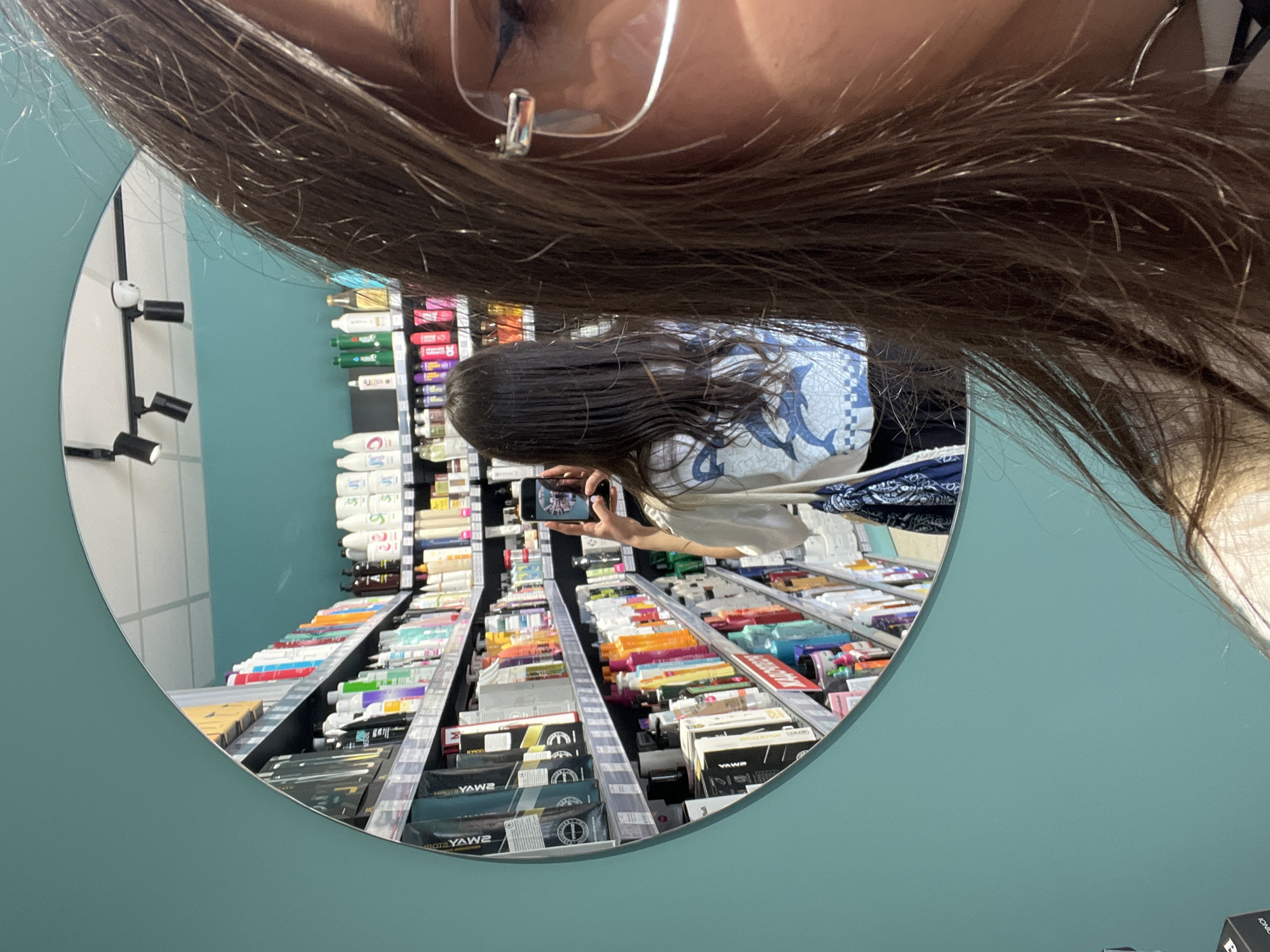 A person with brown hair and glasses is taking a mirror selfie in what appears to be a retail store. The shelves in the background are filled with various beauty and hygiene products. The lighting in the store seems bright and fluorescent. The mirror's frame is light blue. The exact location in Ternopil, Ukraine, is undiscernible.