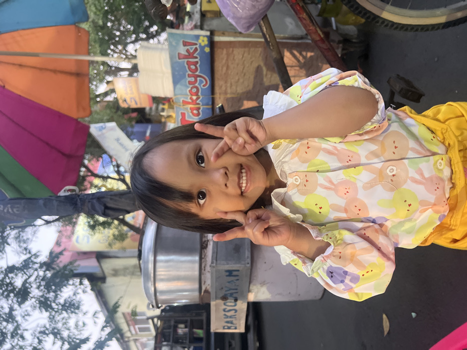 An image captured in Kecamatan Pati, Indonesia, depicts a young child positioned centrally, smiling directly at the camera. The child, with dark hair and bangs, is wearing a long-sleeved, white collared shirt patterned with pastel-colored cartoon rabbit heads and bright yellow lower garments. Both of the child's hands are raised to chest level, each forming a peace sign (V-sign) with outstretched index and middle fingers.
The background reveals an outdoor market or street food vendor scene. To the immediate left of the child is a large, cylindrical, stainless steel steamer pot resting on a white-painted metal stand. A sign attached to this stand displays "BAKSO AYAM" in blue text. Further left, a dark-colored vehicle and parts of buildings are visible. To the immediate right of the child, a large, multi-colored umbrella (segments of pink, green, orange, and purple) provides shade over a vendor stall. Below the umbrella, a white sign with prominent red lettering reads "Takoyaki", accompanied by blue text. A bicycle frame, mostly red-brown, with a purple plastic bag hanging from it, is partially visible on the far right. The scene is illuminated by natural light, indicating daylight hours.