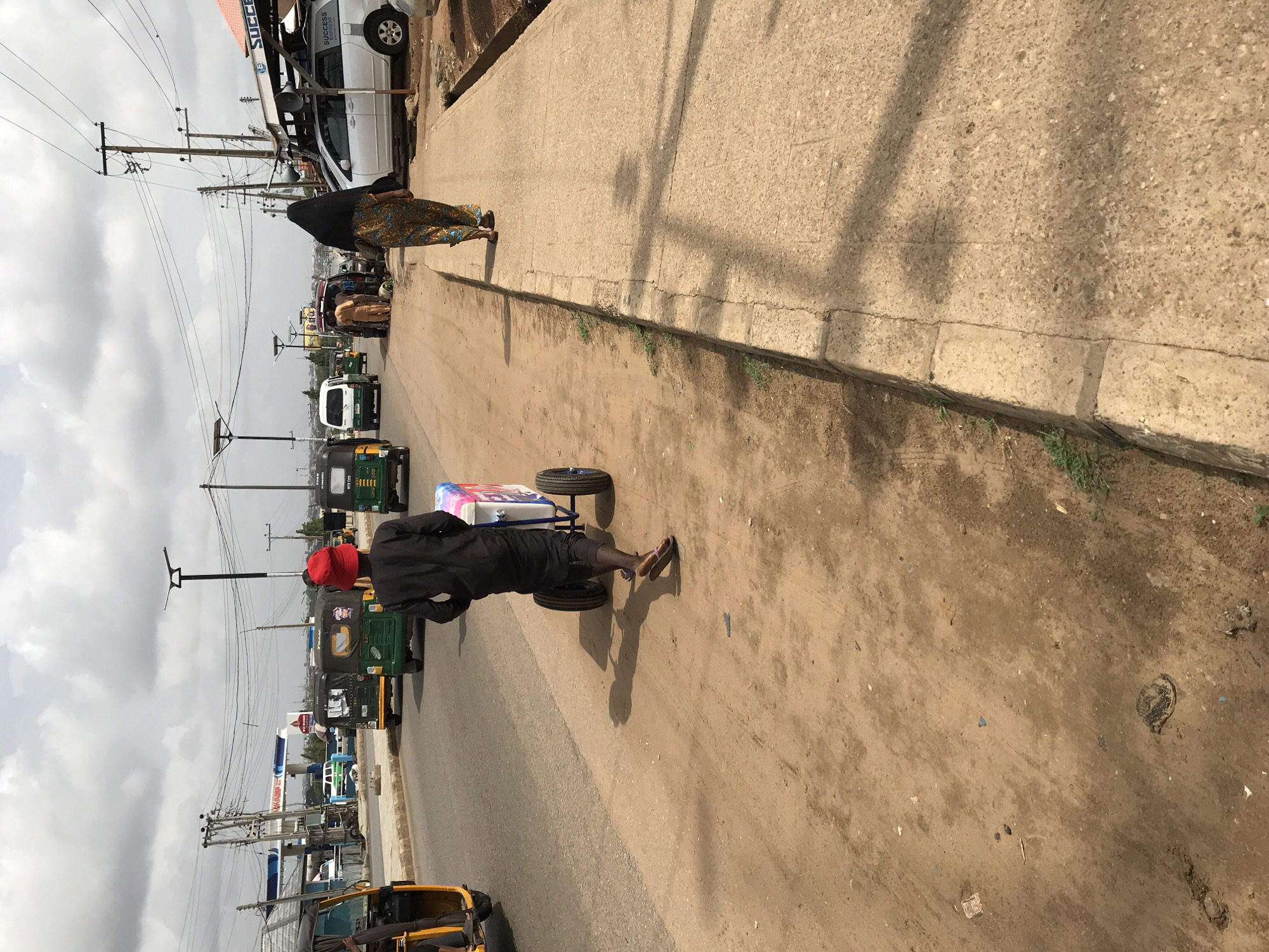 Location: Mandate Estate, Ilorin, Nigeria.
Scene: A street view. A man wearing a red hat and dark clothing pulls a two-wheeled cart with a covered box. The road is partially paved, adjacent to a concrete sidewalk. A woman in traditional attire walks on the sidewalk. Several auto-rickshaws (tuk-tuks) are visible on the road. Utility poles with numerous wires are present. Buildings and commercial signs are visible in the background. The sky is overcast.