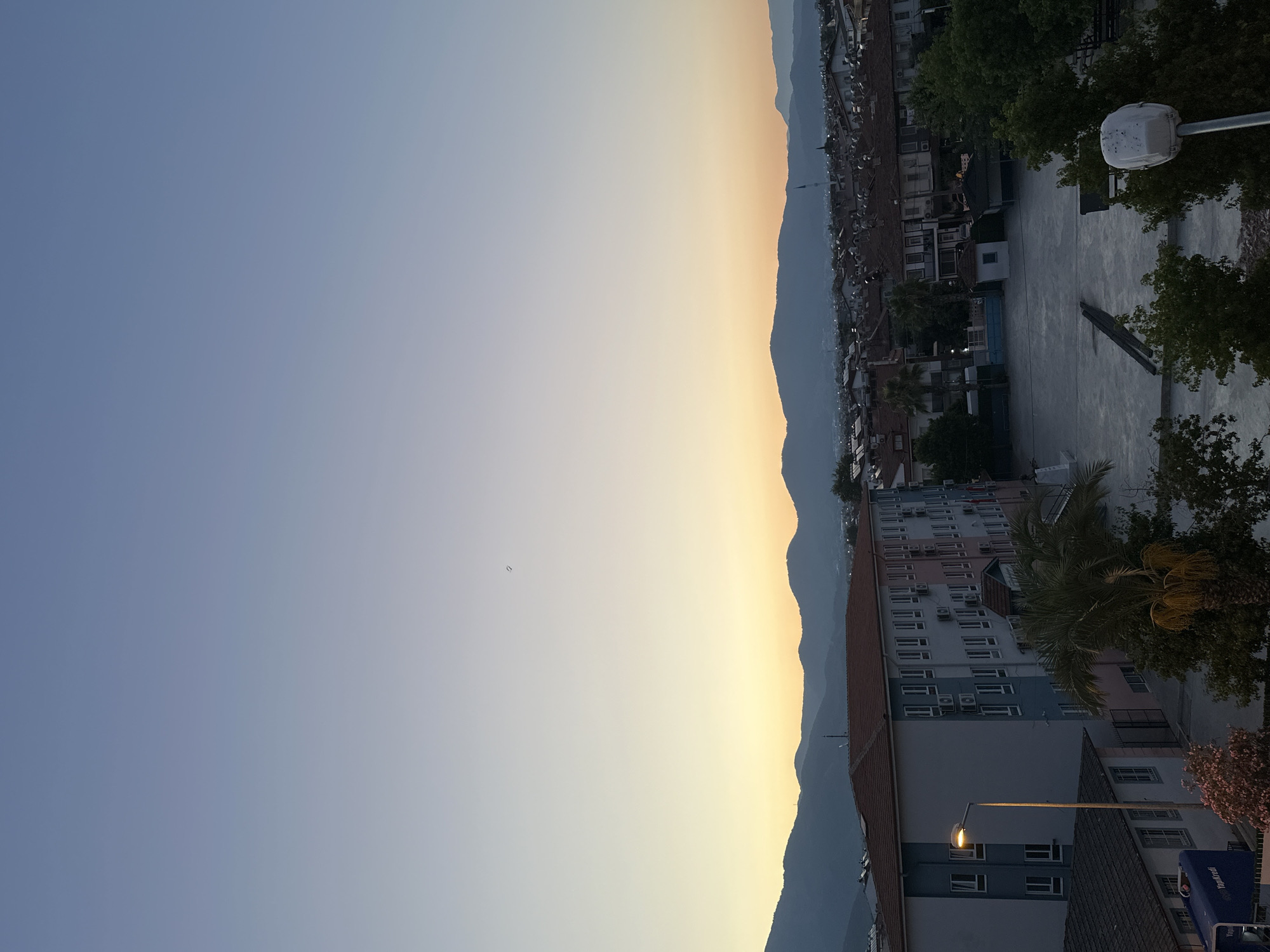 The image shows a wide shot of a city, likely Fethiye, Turkey, at dusk. The sky is a gradient of pale blue to yellow, with mountains silhouetted in the distance. In the foreground, there is a multi-story building with a light-colored facade and rows of windows. Adjacent to it is a large, paved open area, possibly a courtyard or sports field. Several trees, including a prominent palm tree, are scattered throughout the scene. A street lamp stands to the left of the main building, and a white utility box is visible in the lower right corner. The overall atmosphere is calm and serene, suggesting the end of a day.