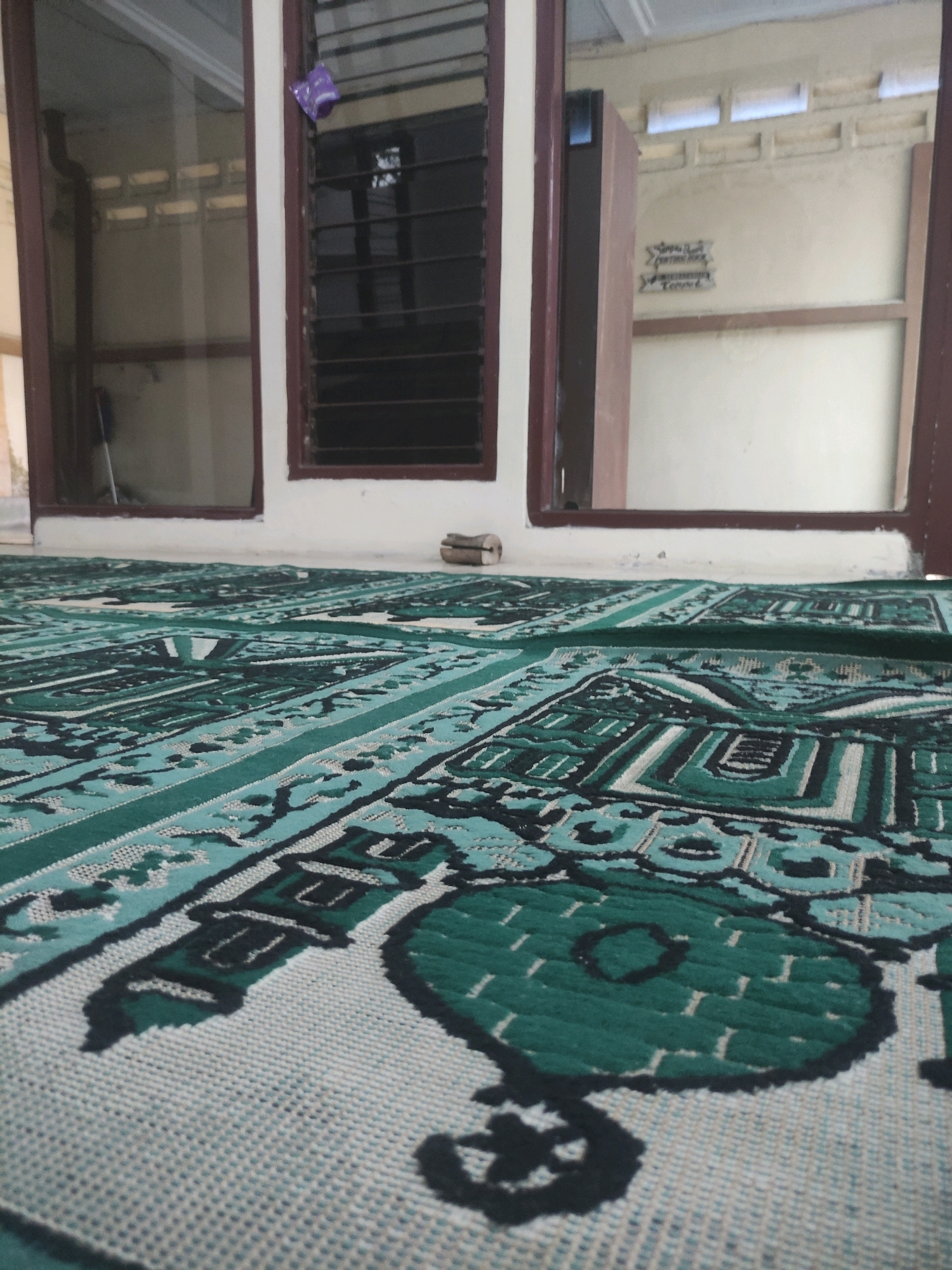 This is a low-angle, close-up shot of a prayer mat with a repeating geometric pattern in shades of green and white. The mat covers the floor of what appears to be a room with large windows. Through the windows, a glimpse of the exterior can be seen, with part of a wall and a doorway visible. There is a small, purple object hanging on the window frame in the background. A thin, rectangular object, possibly a book or a notepad, lies on the windowsill in the center of the frame. The scene is indoors, and the lighting suggests it is daytime. The image appears to have been taken in Ngasem, Indonesia, given the details of the room's construction and the commonality of prayer mats in the region.